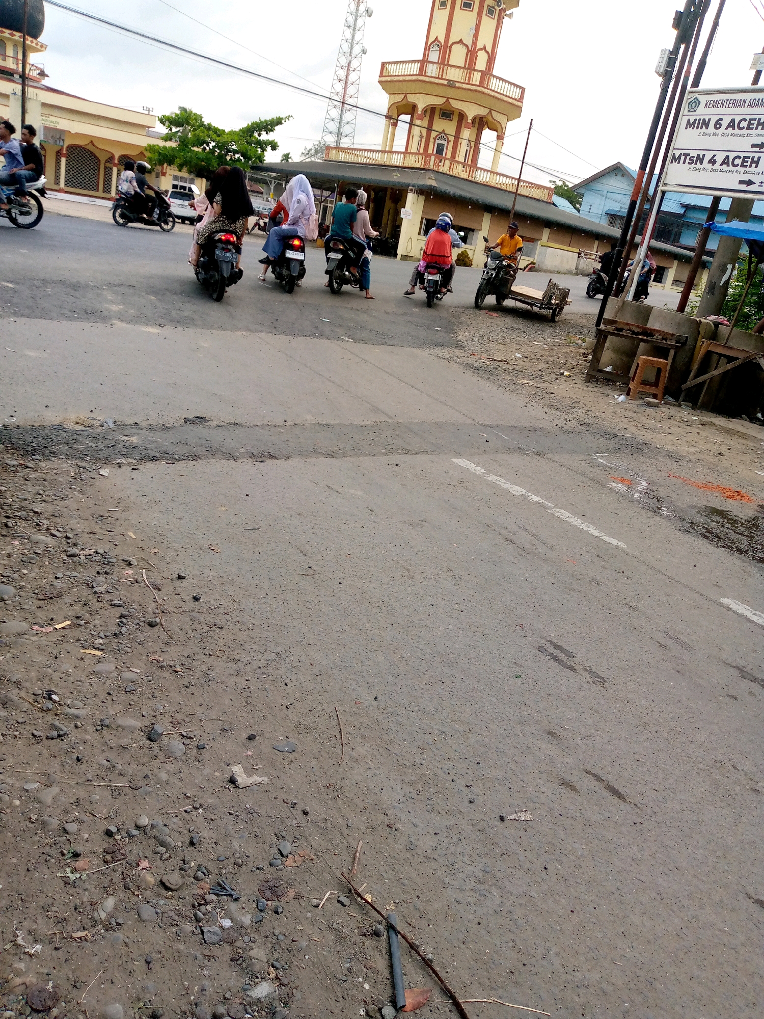 The image depicts a street scene in Keudegeudong, Indonesia, likely on a cloudy day. In the foreground, the asphalt road shows signs of wear and tear, with gravel and debris along the shoulder. Several individuals are riding motorcycles. Two people are on a motorcycle in the left middle ground, one wearing a blue shirt and the other a dark shirt. Further to the right, a motorcycle is occupied by two people, one in a light-colored outfit and the other in a pink and white outfit, both wearing hijabs. Another motorcycle follows, with two people also wearing hijabs. A fourth motorcycle with two passengers, one in a blue shirt and the other in a pink hijab, is also present. On the right side of the street, a motorcycle with a sidecar is ridden by a person in an orange shirt. In the background, a distinctive yellow and red minaret of a mosque stands tall. A telecommunications tower is also visible behind the mosque. Several buildings with light yellow facades line the street. A street sign is affixed to a pole on the right, partially obscured. The environment appears to be an urban or semi-urban setting.