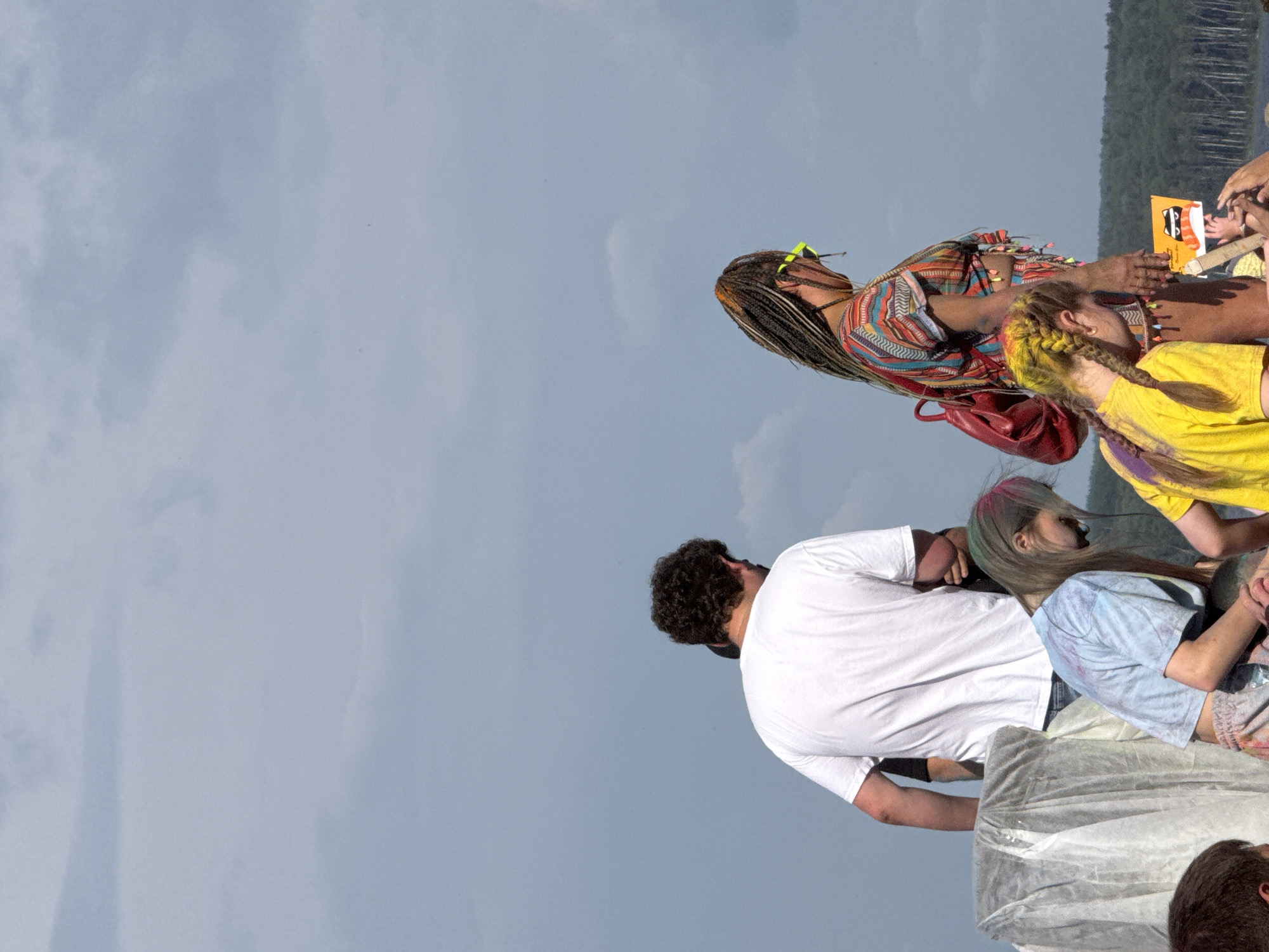 An outdoor medium shot captures a group of people amidst a cloudy sky. In the foreground, three individuals are visible. On the left, a person wearing a light blue tie-dye t-shirt and patterned shorts leans forward. To their right, a person with blonde hair in braids, wearing a yellow t-shirt, also leans forward. Between them, a person with long braided hair and a colorful patterned shirt, wearing bright yellow sunglasses, stands with their back to the camera, holding a red backpack. In the middle ground, a person wearing a white t-shirt stands facing away. The background features a dense line of trees along a body of water under a vast, overcast sky. The overall scene appears to be a gathering, possibly a festival, given the vibrant colors and the presence of holi powder on some individuals.