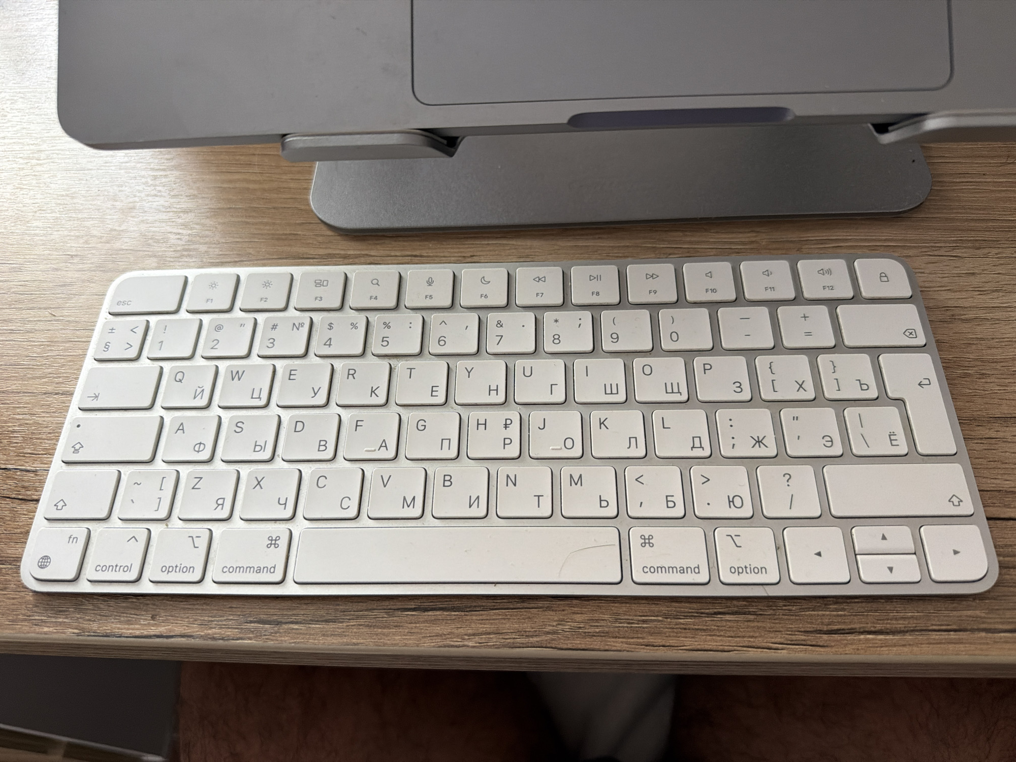 A close-up shot shows an Apple Magic Keyboard resting on a wood-grained surface, likely a desk. The keyboard features Cyrillic characters alongside the standard English alphabet, indicating it is designed for use in a region where the Russian language is prevalent, likely Russia. A portion of a silver-colored Apple monitor or all-in-one computer sits in the upper portion of the frame. The space bar has a crack in it. Below the desk is partially visible the person or persons who took the photo. No specific location in Chaikovsky is discernible from the image.