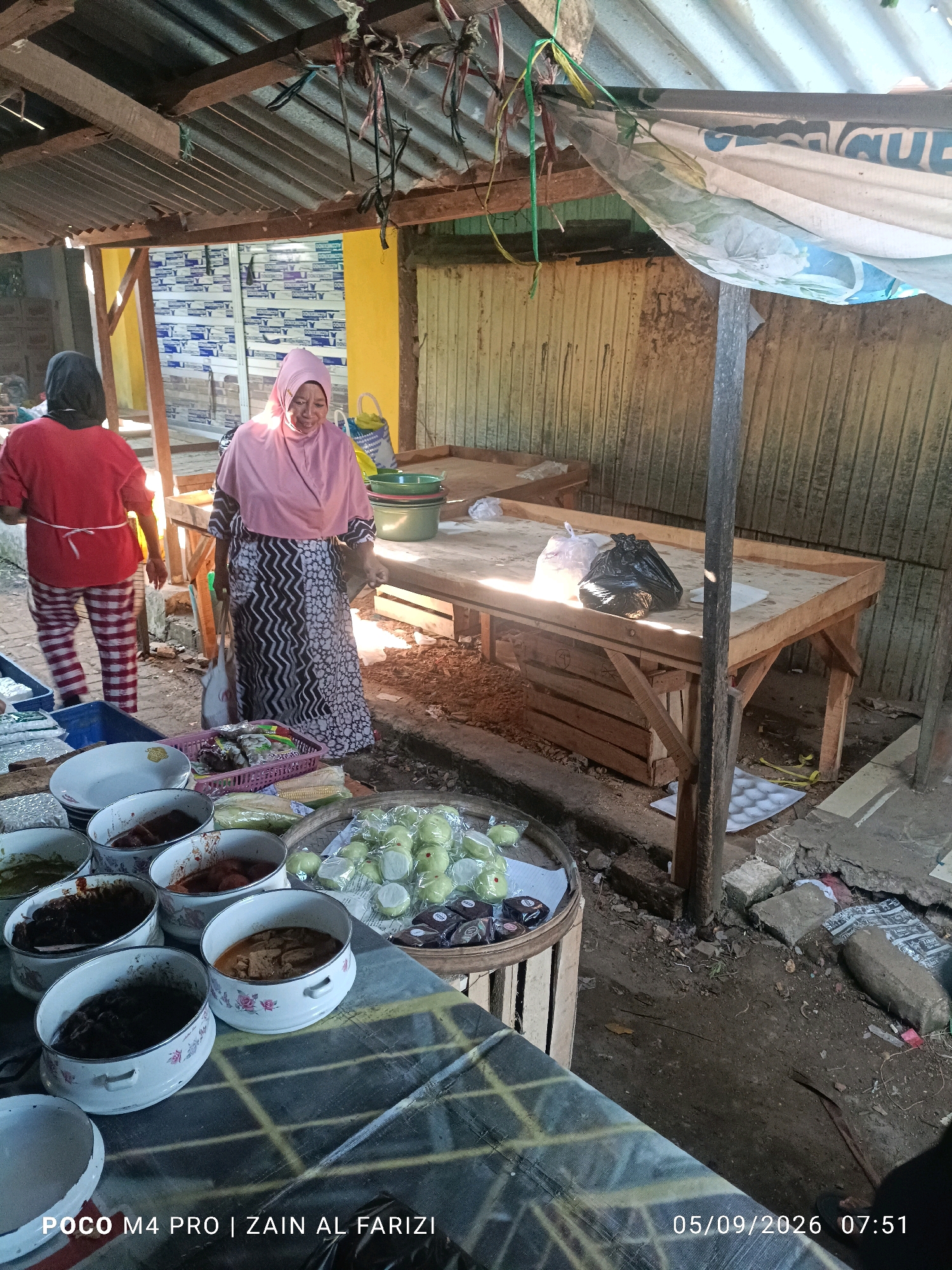 Two women are standing near wooden tables in an outdoor market stall. The woman on the left wears a red shirt and checkered pants, while the woman on the right is wearing a pink hijab and a patterned dress. Several containers of food are displayed on a table in the foreground, including what appear to be small, green fruits and some dark brown pastries. The market stall has a corrugated metal roof supported by wooden beams. The ground is dirt and scattered with debris. The timestamp "05/09/2026 07:51" and text "POCO M4 PRO | ZAIN AL FARIZI" are visible in the lower portion of the image. The location is identified as Arosbaya, Indonesia.
