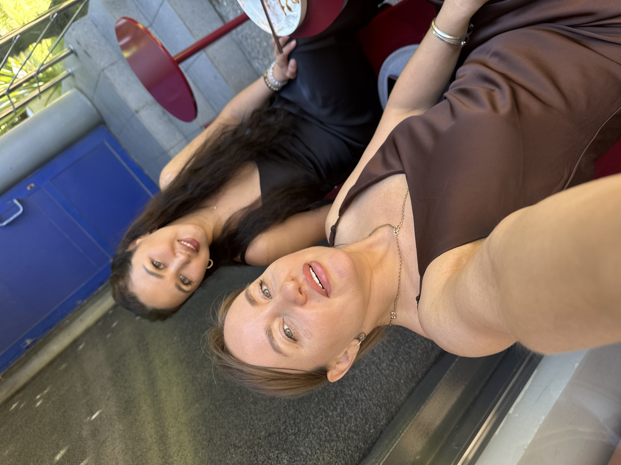 Two women are seated at a table outside, possibly at a cafe or restaurant, with a blue door and railing visible in the background. The woman in the foreground is taking a selfie, wearing a brown satin dress and jewelry. The woman behind her is wearing a black dress and also has jewelry. A red table with a plate of food is also visible. The scene appears to be set in an urban outdoor environment.