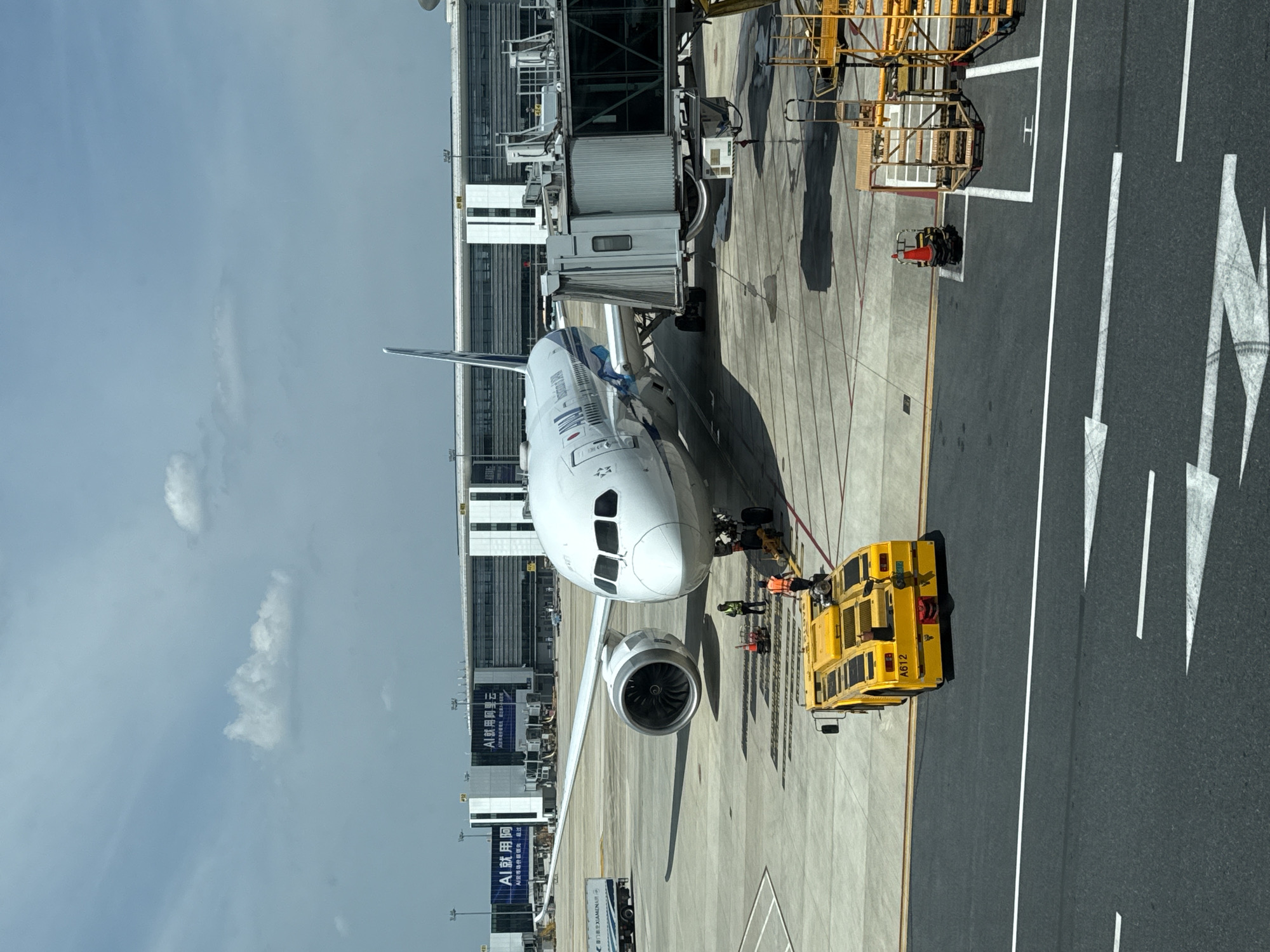 The image depicts an outdoor scene at an airport, likely Hangzhou Xiaoshan International Airport in Xiaoshan District, China, as indicated by signage on the terminal building that reads "AI就用阿..." which translates to "AI use Ah..." followed by characters likely referring to Alibaba's cloud services and "XIAMEN" on a truck. A large, white passenger airplane with blue and white livery is parked at a gate, connected to a jet bridge. The aircraft's front landing gear is visible, and its engines are mounted under the wings.
In the foreground, a yellow pushback tractor is positioned behind the airplane. Several individuals wearing high-visibility vests are working around the aircraft and the tractor, suggesting ground operations are underway. The ground surface is asphalt and concrete, with painted markings for taxiways and parking areas.
In the background, the modern airport terminal building is visible, featuring a glass facade and architectural elements. Signs with Chinese characters are displayed on the terminal. The sky is partly cloudy with a bright, daytime light. Other airport equipment, such as a vehicle and scaffolding, are present in the scene.
