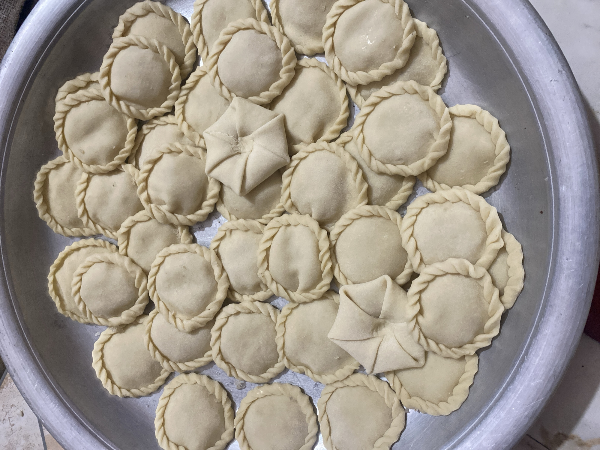 A shallow, circular, metallic tray, likely aluminum, is filled with numerous uncooked, circular pastries. The pastries are arranged in a dense, overlapping pattern across the surface of the tray. Each pastry appears to be made of a light-colored dough, possibly flour-based, and is crimped along the outer edge with a decorative, braided pattern. Several pastries are plain circles, while others have a more folded or star-shaped appearance in the center, suggesting different filling methods or shapes. The tray itself has a slightly worn, brushed metal finish and rests on a surface that appears to be a tiled floor. The lighting is overhead, creating soft shadows and highlighting the texture of the pastries and the tray. This scene likely represents food preparation, possibly for a special occasion or sale, within a domestic setting in Dhaka, Bangladesh, given the commonality of such pastries and preparation methods in the region.
