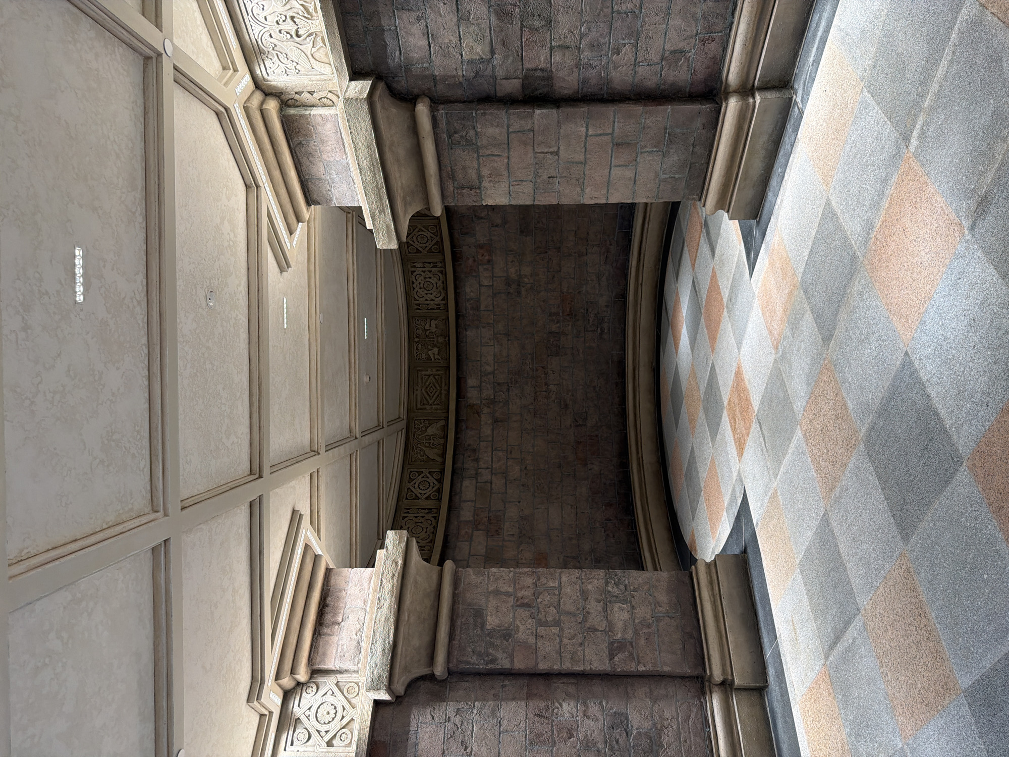 The image depicts a circular courtyard or atrium with an octagonal opening in the foreground, likely a public space within Đà Nẵng, Vietnam. Two large, stone-clad columns, constructed from dark bricks with ornate stone capitals, frame the entrance to the circular area. The floor of the courtyard is paved with a checkerboard pattern of grey and peach-colored granite tiles. The curved wall of the courtyard is also made of dark bricks and features decorative friezes with geometric patterns and stylized animal motifs near the ceiling. The ceiling itself is a coffered design with a textured, off-white finish and recessed lighting. No people are visible in the image. The environment appears to be an indoor or covered architectural space, possibly part of a museum, historical building, or a themed area within a larger complex.