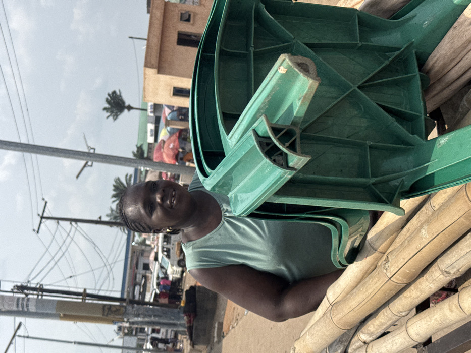 An outdoor, daylight scene in a bustling commercial area of Ogbaku, Nigeria, is depicted under a partly cloudy sky. In the immediate foreground, an adult female with dark skin, braided hair, wearing a light green sleeveless top and a gold hoop earring, smiles directly at the viewer. She is positioned behind a stack of green plastic chairs, one of which is prominently inverted, revealing its structural underside. Below and to the left of the chairs are several bundled bamboo poles. The mid-ground reveals a busy street with multiple white passenger vans and other vehicles, indicating a thoroughfare or market hub. A petrol station with a blue canopy is visible on the left side of the street. Numerous individuals are present among market stalls featuring colorful red and orange umbrellas. The background includes utility poles with an array of overhead wires on the left, a light brown multi-story building on the right, and scattered palm trees.