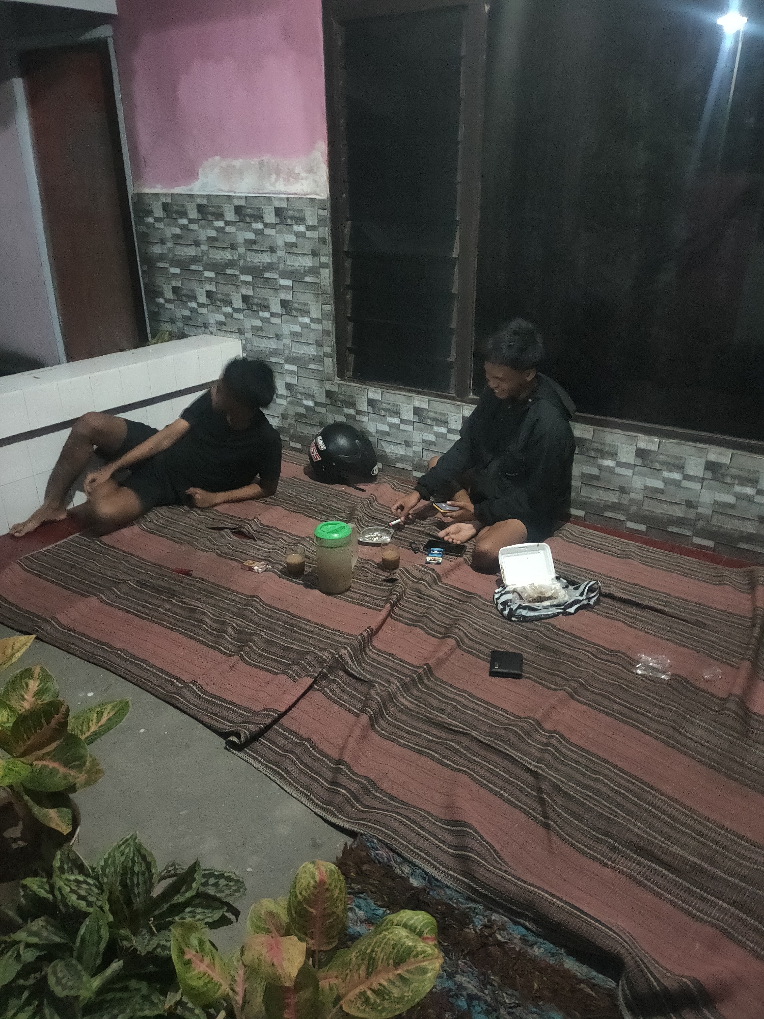Two young men are on a woven mat on the ground, likely in an outdoor patio area. The man on the left is lying down wearing a black t-shirt and shorts, with his legs bent. The man on the right is sitting up wearing a black hooded jacket and shorts, holding a cigarette and looking at his phone. There are several items on the mat, including a helmet, a green plastic container, cigarette packs, a white food container with food inside, a wallet, and some plastic wrappers. In the foreground, there are several potted plants with green and pink variegated leaves. The background shows a wall with patterned tiles and a pink painted section. The overall scene suggests a casual gathering in the evening.