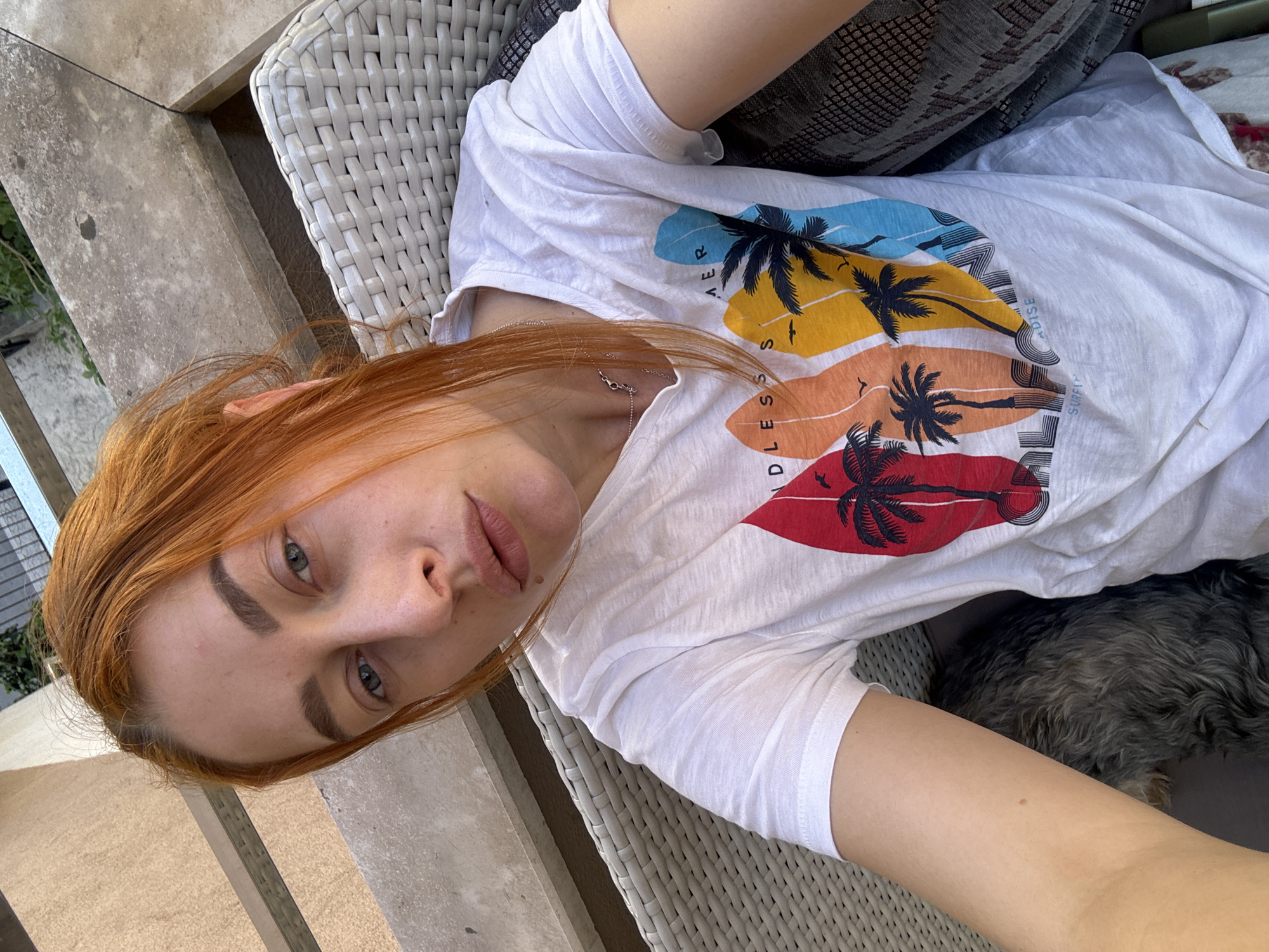 A woman with red hair and fair skin is lying on a white wicker chair, wearing a white t-shirt with a graphic design of palm trees on surfboards with the words "ENDLESS SUMMER PARADISE SURFING". She is looking directly at the camera. A small dog is lying at her side. The location is outdoors, likely a patio, based on the exterior wall, vegetation, and the type of chair. While a specific location in Budva, Montenegro cannot be determined from the image, the exterior environment suggests it could be there.