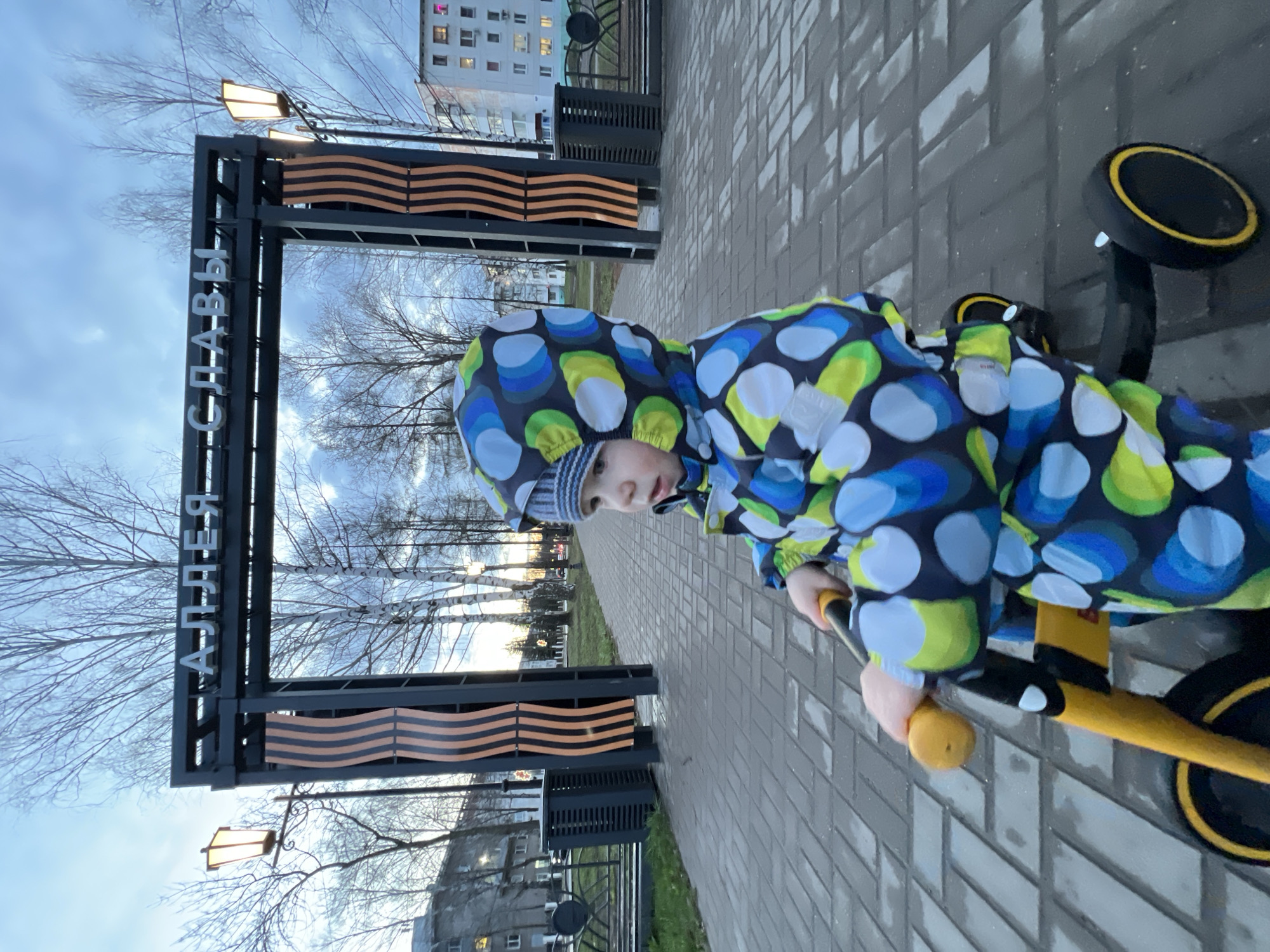 A low-angle, eye-level shot captures a young child, likely a boy, dressed in a vibrant blue, green, and yellow patterned hooded winter jacket and a striped hat. He is seated on a yellow tricycle with black wheels, holding the handlebars, and looking directly towards the camera.
The setting is an outdoor paved area, possibly a park or alley. In the background, a prominent black metal archway or gate structure is visible. White Cyrillic letters on the archway spell out "АЛЛЕЯ СЛАВЫ," which translates to "Alley of Glory." Below the horizontal beam of the archway, there is a decorative element resembling a wavy black and orange striped ribbon, characteristic of the Ribbon of Saint George. Bare trees with thin branches are visible behind the archway, suggesting late autumn, winter, or early spring. Distant residential buildings are also present under a muted, overcast sky. The ground is paved with grey interlocking bricks, appearing slightly damp. The lighting suggests it is late afternoon or early evening, with a soft, ambient glow. The overall atmosphere is calm and cool.