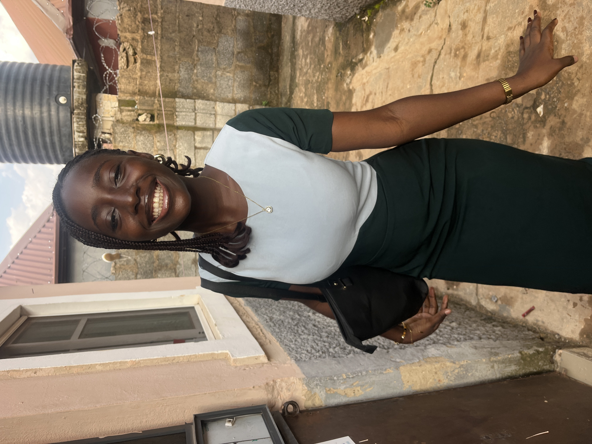 A young Black woman with braided hair is smiling at the camera in what appears to be an outdoor setting in Abuja, Nigeria. She is wearing a two-toned dress with a light blue top and a dark green skirt. She is carrying a black handbag over her left shoulder and is wearing a gold necklace and a gold bracelet. Behind her, there is a wall made of stone and brick, a water tank on top of a building, and barbed wire. The ground is a light brown, dusty earth. To her left, there is a building with a cream-colored exterior and a window.