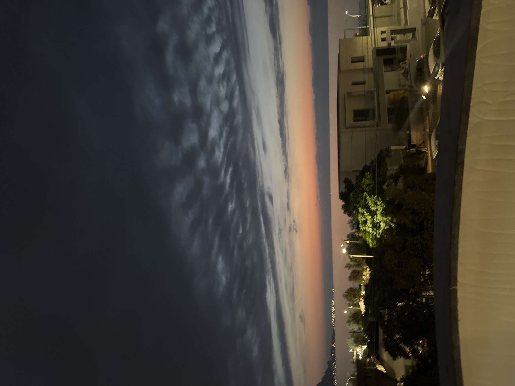 This is a nighttime aerial photograph of a coastal city, likely Capaci, Italy, taken at dusk. A dramatic sky dominates the upper portion of the frame, filled with dark, textured clouds that stretch horizontally across the image. Below the clouds, a gradient of color transitions from a soft orange at the horizon to a pale yellow, suggesting the last remnants of sunset. The sea is visible in the mid-ground, with a distant, illuminated coastline. Several streetlights dot the landscape along the shore, casting a warm glow. In the foreground, modern, light-colored buildings are visible, with some trees and foliage interspersed. Several cars are parked or driving on a street in front of the buildings, their headlights briefly illuminating the scene. The foreground also features a white, curved structure, possibly a railing or balcony, partially obscuring the view of the city below. The overall impression is one of a peaceful, urban coastal setting transitioning into night.