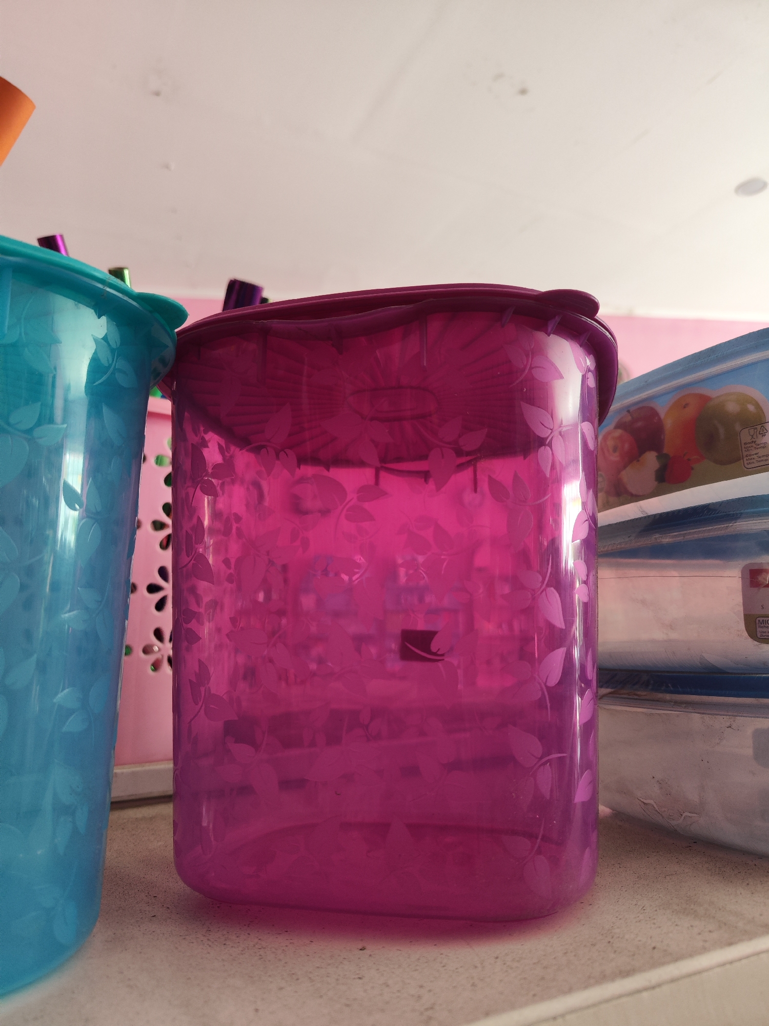This is a close-up, eye-level, indoor, medium shot of plastic food storage containers, taken on a speckled countertop. In the foreground and slightly to the left, a bright blue, translucent plastic container with a leaf pattern is visible. To its right, a larger, vibrant pink, translucent plastic container with a similar leaf pattern dominates the center of the frame. Both containers appear to be on a shelf. Behind the pink container, to the right, are stacked clear plastic food containers with blue labels depicting apples. The background consists of a light-colored ceiling with some subtle markings and a pale pink wall. The lighting suggests an ambient indoor environment. No people or actions are depicted. The location in Sragen, Indonesia cannot be determined from the visual information.