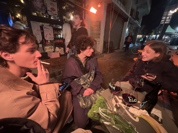 Three young adults sit at an outdoor cafe table at night. A man on the left smokes a cigarette while looking to the side. Another person sits in the middle, looking towards the woman on the right, who holds a phone. Wine glasses with names written on them and a wine bottle are on the table, along with a bundle of green onions or leeks. A bicycle and other pedestrians are visible in the background, along with a bar display inside the establishment.