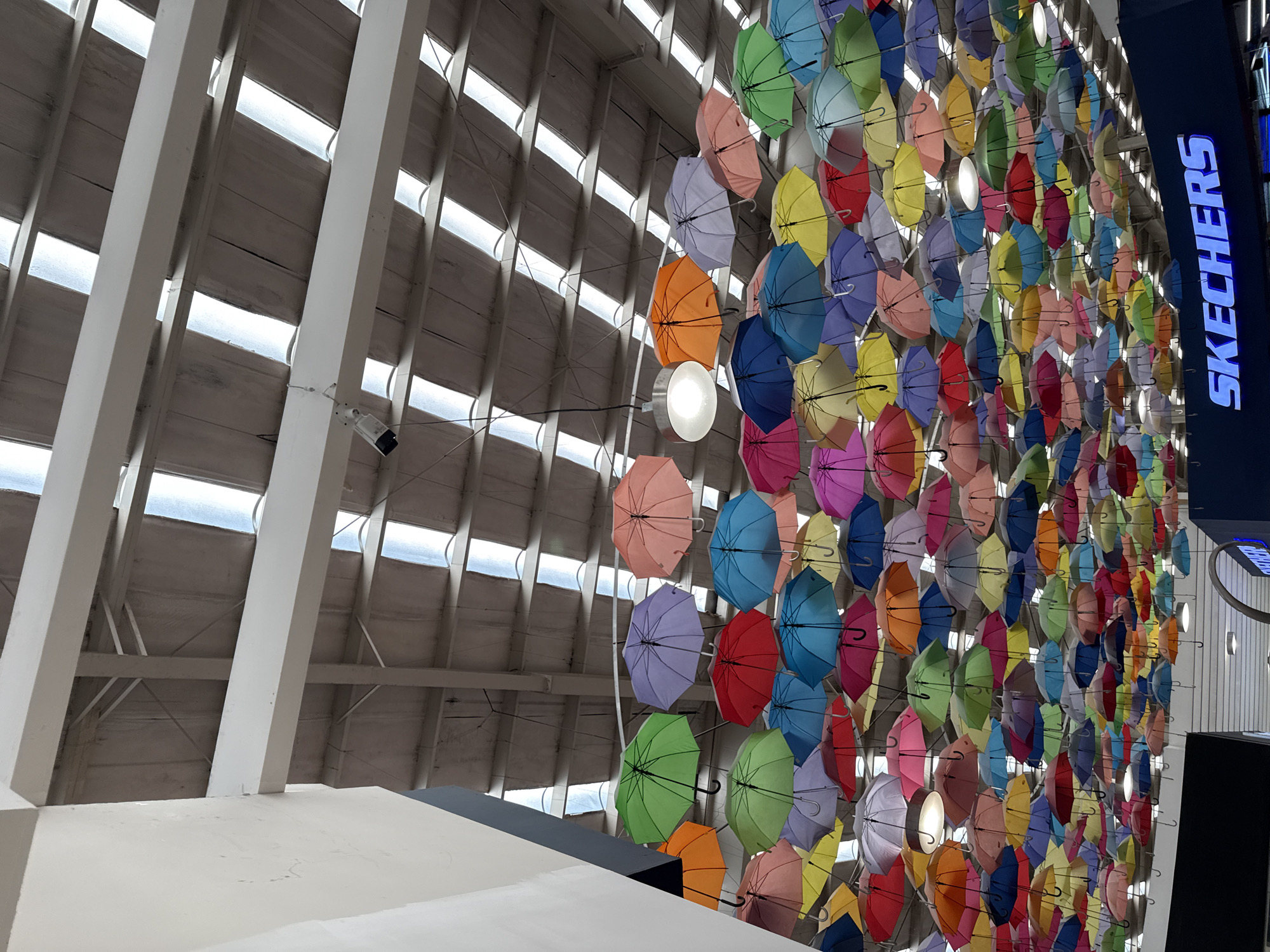 The image shows an interior space with a decorative display of colorful umbrellas attached to a wall. A "SKECHERS" sign is visible at the top right. The space has a high, angled ceiling with linear architectural elements and some translucent panels. The overall impression is of a retail or commercial environment, possibly a shopping mall or department store. The location in San Francisco Ocotlán, Mexico is not explicitly evident from the image.