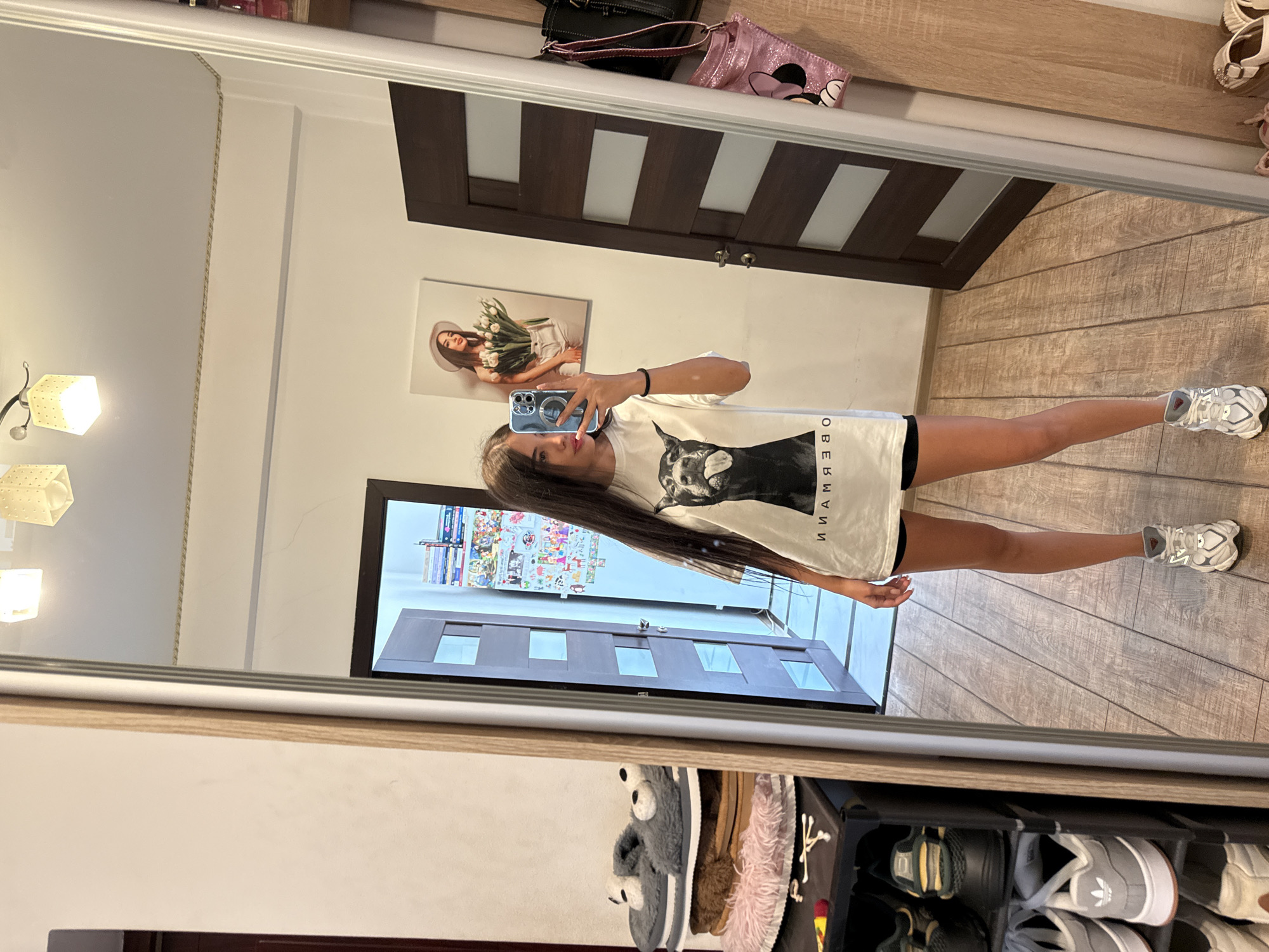The image shows a young woman with long brown hair taking a mirror selfie in what appears to be a bedroom or dressing area. She is wearing a white t-shirt with a black graphic of a dog and black shorts. She has on white athletic shoes. In the background, a shoe rack is visible to the left, holding various pairs of shoes and slippers. A large mirror dominates the right side of the frame, reflecting the scene. Behind the woman, a dark wooden door with glass panels is visible. On the wall, a framed picture of a woman holding tulips hangs. The floor is made of wooden planks. To the far right, a closet door is partially visible, with a pink bag with a Mickey Mouse design hanging on it. The overall impression is a casual, home environment. There are no discernible landmarks that would indicate a specific location within Chișinău, Moldova.