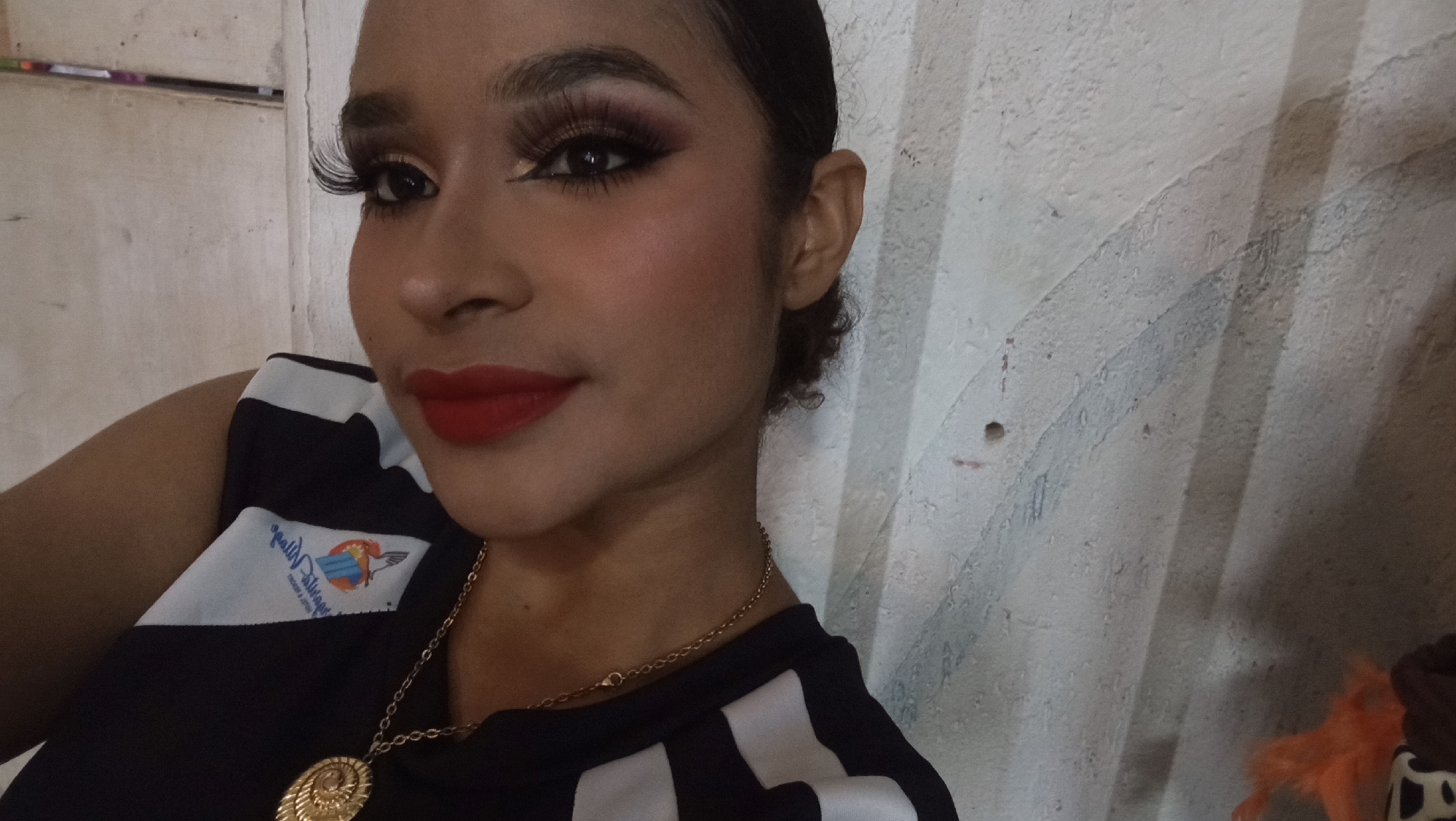 This image depicts a young woman in a close-up, head-and-shoulders shot, positioned against a light-colored, vertically grooved wall, in Porlamar, Venezuela. The woman has a medium-brown skin tone and dark hair pulled back, with small curls visible near her right ear. She is wearing heavy makeup, including prominent false eyelashes, dark winged eyeliner, defined eyebrows, blush on her cheeks, and bright red lipstick. She looks slightly to the viewer's right with a soft, closed-mouth smile. Her attire consists of a black sleeveless top with broad white horizontal stripes. On the left side of her top, a partial, mirrored logo is visible, featuring text that appears to be "Molly's Dream" and a graphic element with blue and orange colors, possibly depicting a bird or an abstract design. She wears a gold-colored chain necklace with a large, circular gold pendant featuring an intricate spiral or shell pattern. The background wall is textured with vertical ridges and shows some faint greyish-blue streaks or marks. A small, dark hole is also visible on the wall to the far left. A small, out-of-focus detail of orange fluffy material and a brown object with white spots is barely discernible in the bottom right corner.