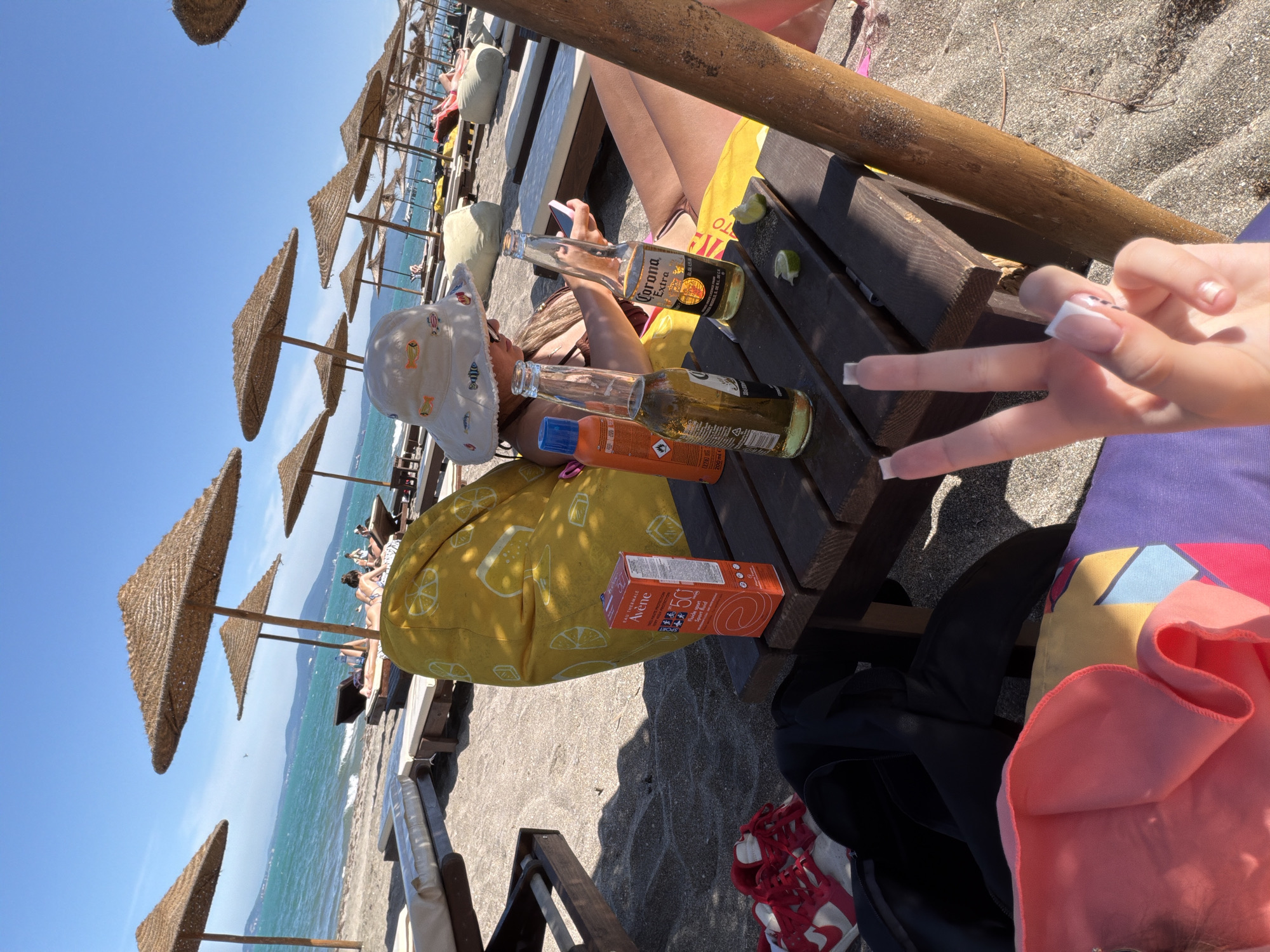 This is a photograph taken on a sunny day at the beach in Burgas, Bulgaria. The scene depicts a leisure setting with multiple lounge chairs and straw umbrellas set up on the sand. In the foreground, a person is seated on a yellow beanbag chair, wearing a white hat with fish motifs. In front of them is a wooden table with two bottles of Corona beer, two lime wedges, a can of sunscreen, and a cell phone. To the left of the table are a pair of red and white sneakers and a black bag. Another person is visible in the background, sunbathing on a lounge chair. The sea is visible in the distance, with a clear blue sky overhead. The overall atmosphere is one of relaxation and enjoyment of a summer day at the beach.