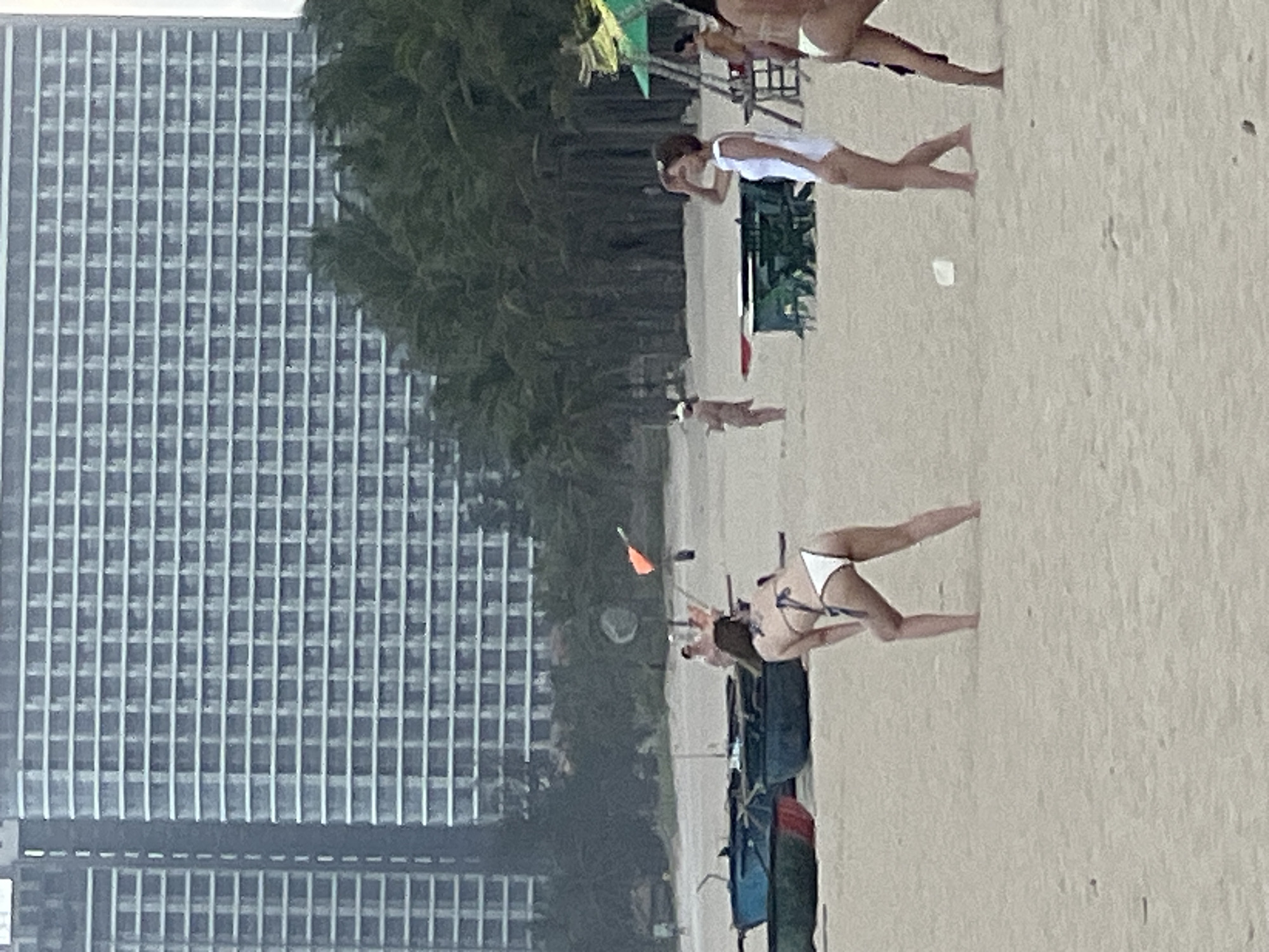The image depicts a beach scene with several people visible in the foreground and mid-ground, set against a backdrop of a large, modern building and palm trees. In the foreground, a woman in a white bikini is bent over, as if preparing to play a sport. To her right, another woman in a white one-piece swimsuit is standing and looking to her left, with her hand shielding her eyes. Further to the right, part of a third woman's legs are visible. In the mid-ground, there are several small boats on the sand and a table with chairs. In the background, a multi-story building with a grid-like facade dominates the skyline, with palm trees in front of it. The overall atmosphere suggests a casual beach day in a resort area, likely in the city of Đà Nẵng, Vietnam, known for its long sandy beaches and modern high-rise hotels. The building in the background is characteristic of hotels found along Đà Nẵng's coastline.