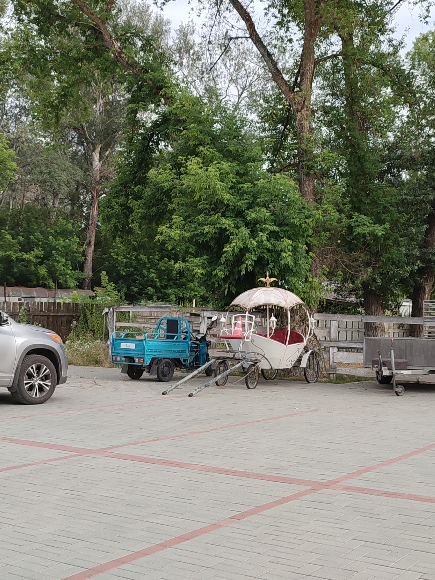 A light-colored car is parked in a lot with a light gray paver surface marked with red lines. To the right of the car is a small blue truck. Further to the right is a white carriage with a decorative canopy and red seating. In the background, there is a wooden fence and numerous trees. The location is identified as Shcherbani, Ukraine.
