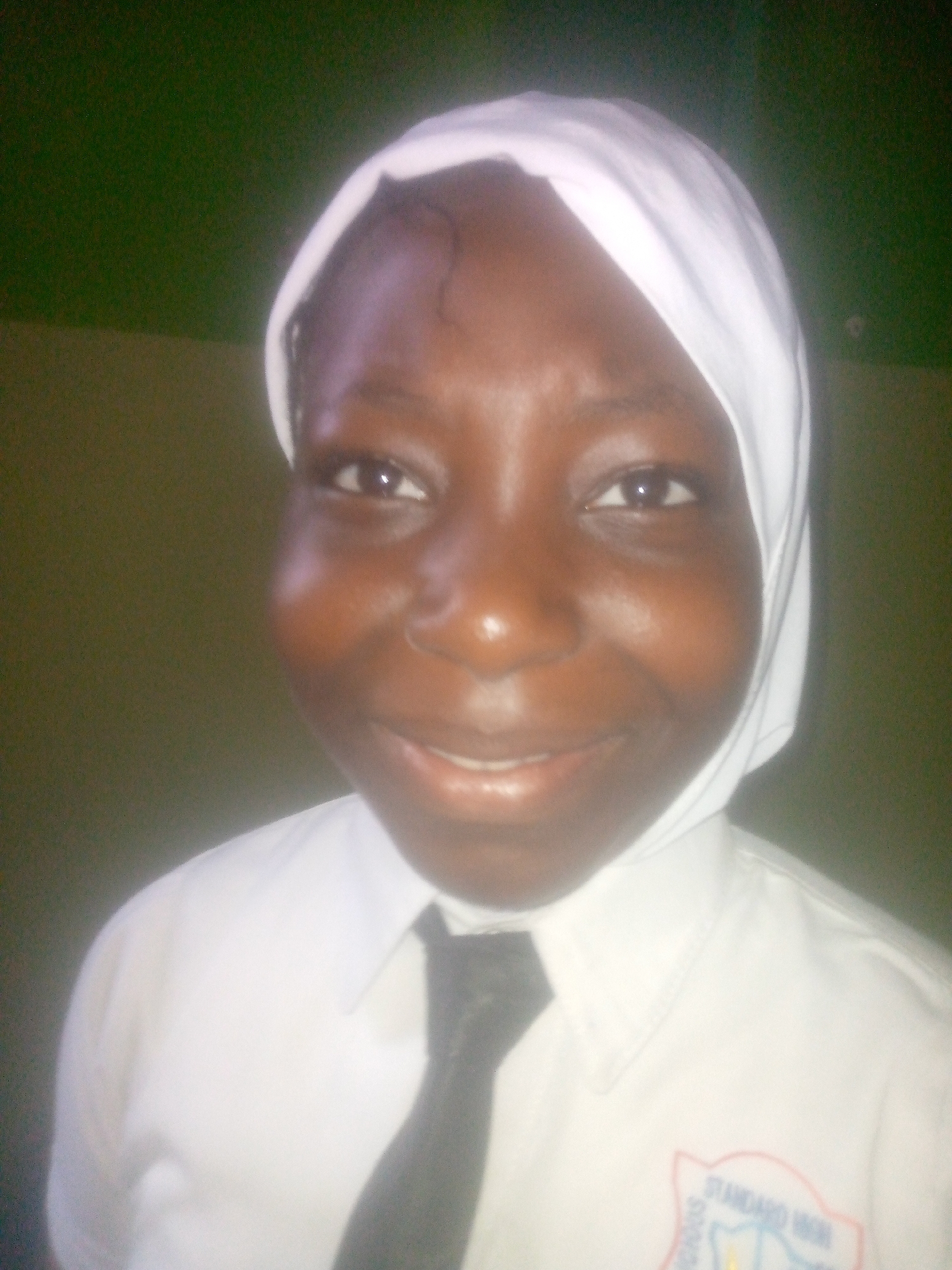 A young, dark-skinned person is smiling and looking directly at the camera. The person is wearing a white shirt with a black tie and a white head covering. A patch on the shirt reads "STANDARD HIGH SCHOOLS". The background is a blurred, dark green color. The location is likely Ikorodu, Nigeria, based on the patch on the shirt indicating the school is located there.