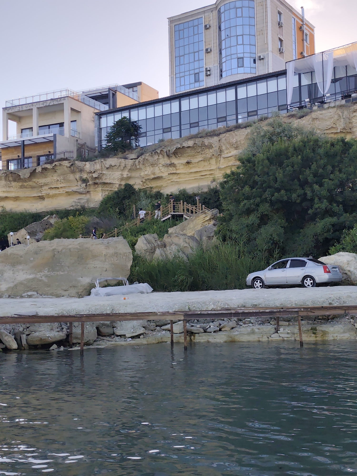 The image depicts a coastal scene in Aktau, Kazakhstan, likely during late afternoon or early evening, as suggested by the soft, diffused light. The setting is characterized by a series of modern buildings perched on a rocky, layered cliff face overlooking a body of calm, dark water.
In the foreground, the water's surface is gently rippled, with some reflections catching the light. A weathered wooden structure, possibly a pier or a walkway, runs along the edge of the water, supported by metal posts. Beyond this structure, a sandy or gravelly area leads up to the base of the cliffs, where large rocks and dense green vegetation are interspersed with the sedimentary rock formations.
On the cliff face, a wooden staircase with railings winds its way up, indicating a path for access to the buildings above. Several people are visible on the stairs and along the cliff, some casually walking, others appearing to be interacting or observing the surroundings. One person is dressed in white, another in dark clothing.
Dominating the upper portion of the image are contemporary buildings with large glass facades, reflecting the sky and surrounding environment. These structures appear to be hotels or upscale residences, exhibiting a modern architectural style. One building features a curved, glass-fronted section, while another has a more angular design with balconies and terraces. A white canopy or awning is visible on one of the buildings.
To the right, a silver sedan is parked on the gravelly area near the base of the cliff, adjacent to thick green foliage and a large boulder. A partially covered object, possibly a boat or equipment, lies on the ground near another large rock formation in the mid-ground.
The overall impression is one of a developed coastal area that blends modern architecture with the natural ruggedness of the cliffs and sea. The atmosphere seems relaxed and peaceful. There is some faint, illegible text on the large rock in the mid-ground.