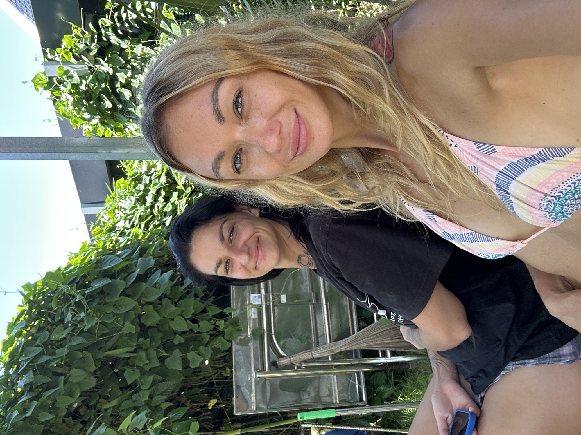 Two women are sitting outdoors in Mũi Né, Vietnam. The woman on the left has dark hair and is wearing a black t-shirt and plaid shorts. She is holding a blue cell phone in her left hand. The woman on the right has blonde hair and is wearing a pink and white striped bikini top. Both women are smiling. Behind them, there is lush green foliage and a metal structure that appears to be part of an outdoor seating area. A broom and a green tool are leaning against the metal structure. The sky is clear and blue.