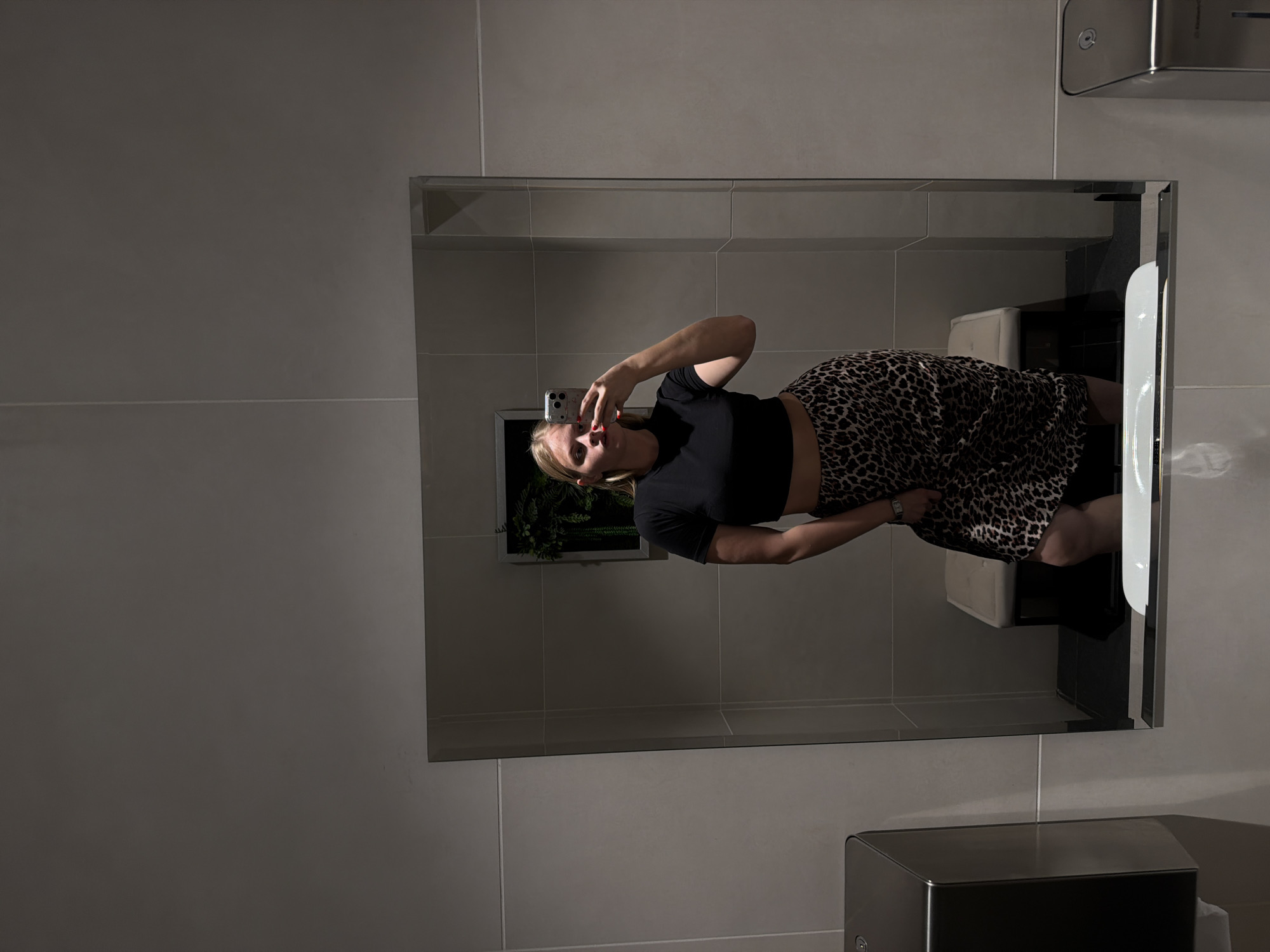 A woman stands in front of a mirror taking a photo with her cell phone. She is wearing a black short-sleeved crop top and a leopard print skirt. A plant is visible in the background, reflected in the mirror. The surrounding environment appears to be a tiled bathroom, containing a hand dryer and paper towel dispenser. The location in Izhevsk, Russia is unknown.