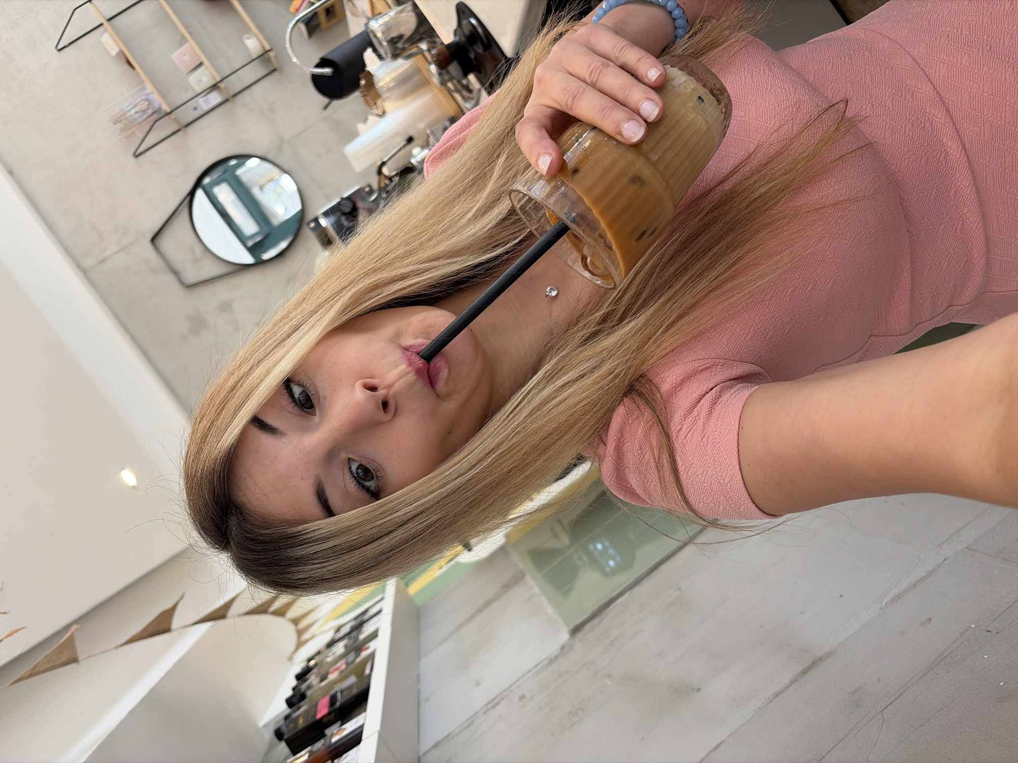 A woman with long blonde hair and a pink dress is holding a glass of iced coffee with a black straw and drinking from it. She is indoors, possibly in a cafe. In the background, there are shelves with various items, a mirror, and a coffee machine. Pennants are hanging in the corner. The location could be Budva, Montenegro, although this is not definitively ascertainable from the image alone.