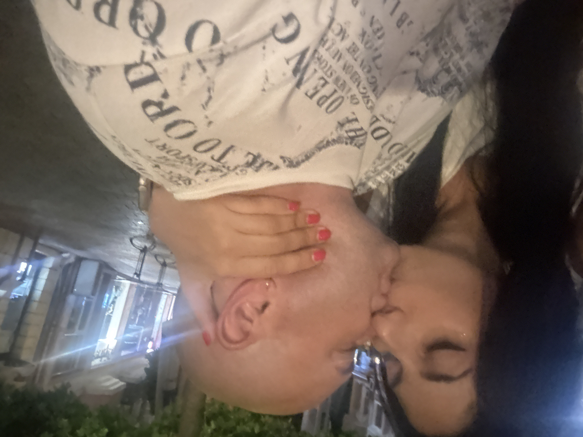 A close-up, medium shot shows a bald man and a woman in an intimate embrace, their lips touching. The man, seen from the side, has a hand on the back of his head, with bright red nail polish on his fingernails. The woman, on the left, has dark hair and is leaning in to kiss him. She is wearing a white top. The man is wearing a white t-shirt with text and graphics printed on it. The background is blurred and appears to be an outdoor urban setting at night, possibly a street in Plovdiv, Bulgaria, with storefronts and trees visible. The lighting is dim and warm.