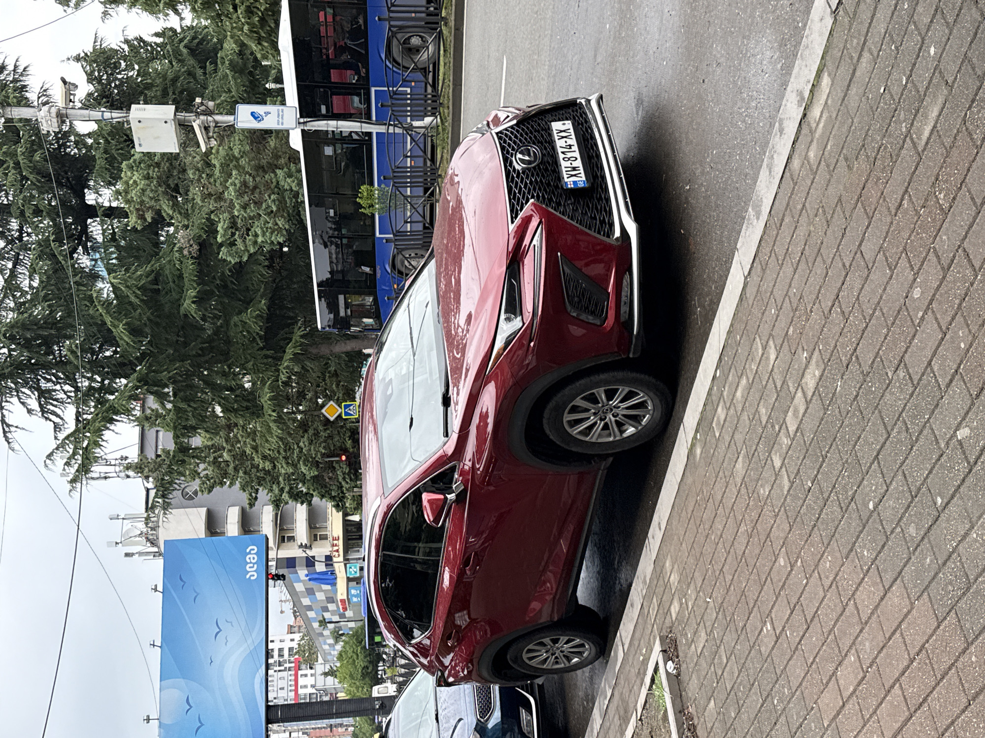 An exterior medium shot captures a parked red Lexus NX 300 SUV on a wet, brick-paved sidewalk next to a road. The vehicle is angled towards the viewer, with its front on the right side of the frame. A license plate on the front of the car reads "XW-814-XX". A dark blue SUV is partially visible to the left of the Lexus. In the background, a blue bus with black-tinted windows is driving on the road. Trees line the street, and beyond them, buildings and a billboard displaying a sky scene with birds are visible. Traffic lights, utility poles with cameras, and street signs are also present. The sky is overcast.