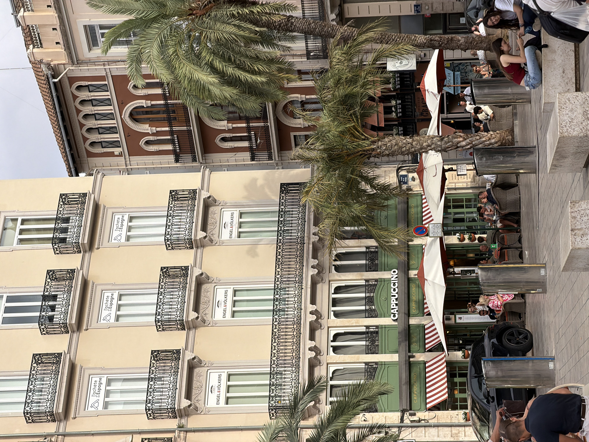 The image depicts an outdoor seating area in front of a building with a cafe named "CAPPUCCINO". Several people are seated at tables under umbrellas, some engaged in conversation and others looking at their phones. A black SUV is parked to the left of the frame. Palm trees are interspersed throughout the scene, indicating a warm climate. The architecture suggests an urban environment with a mix of older and newer buildings. One of the buildings has balconies with ornate railings. The sky is overcast.