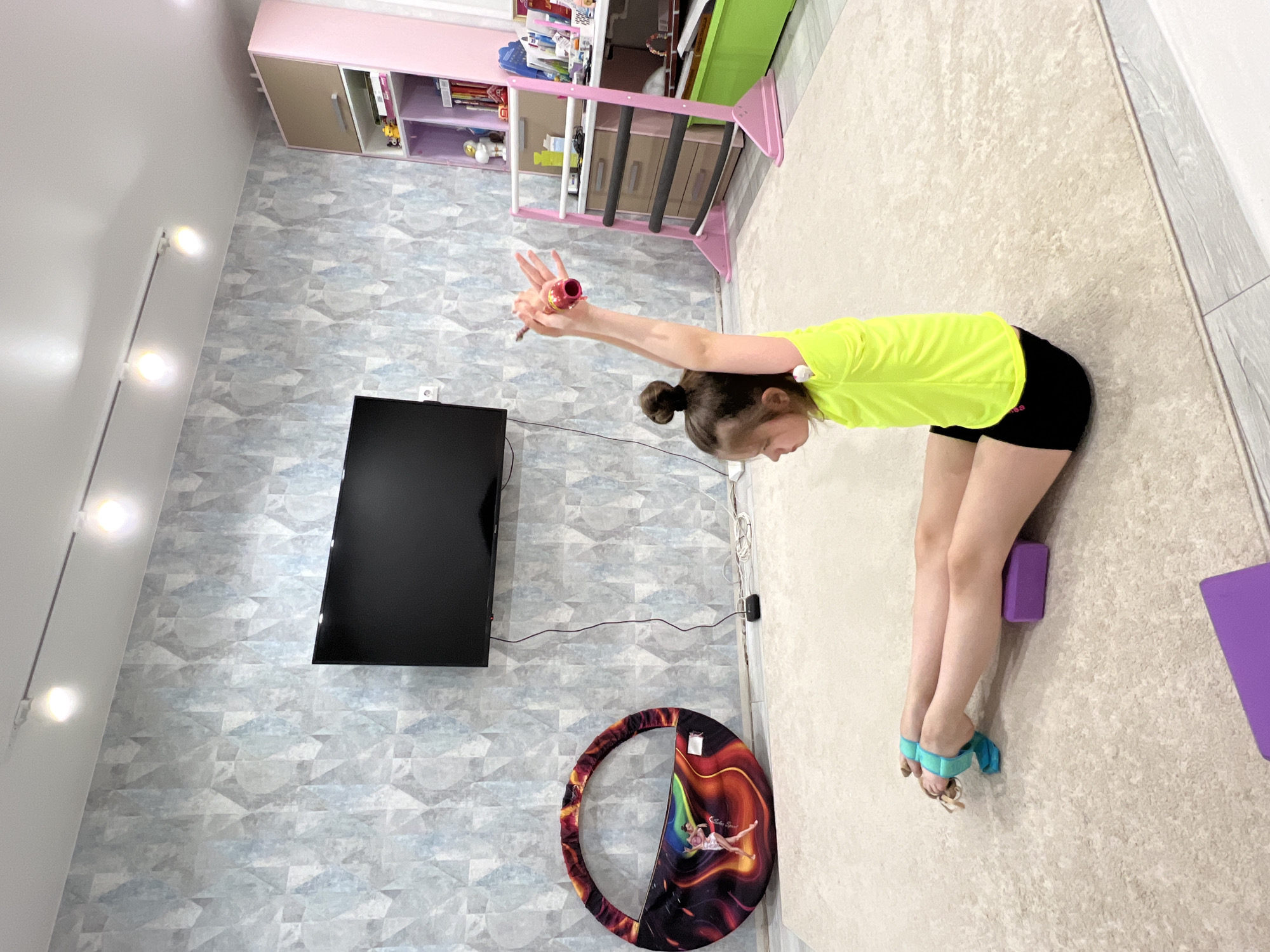 A young girl in a neon yellow shirt and black shorts is performing a gymnastic stretch in a room in Chaikovsky, Russia. Her arms are extended overhead holding a pink and red gymnastic club. She is standing on a beige carpet, using a purple yoga block for support under her thighs and her ankles are secured with teal-colored straps. The room contains a TV mounted on a blue patterned wallpaper, a children's climbing frame, a storage unit, and a gymnastic hoop. The lighting in the room is bright.