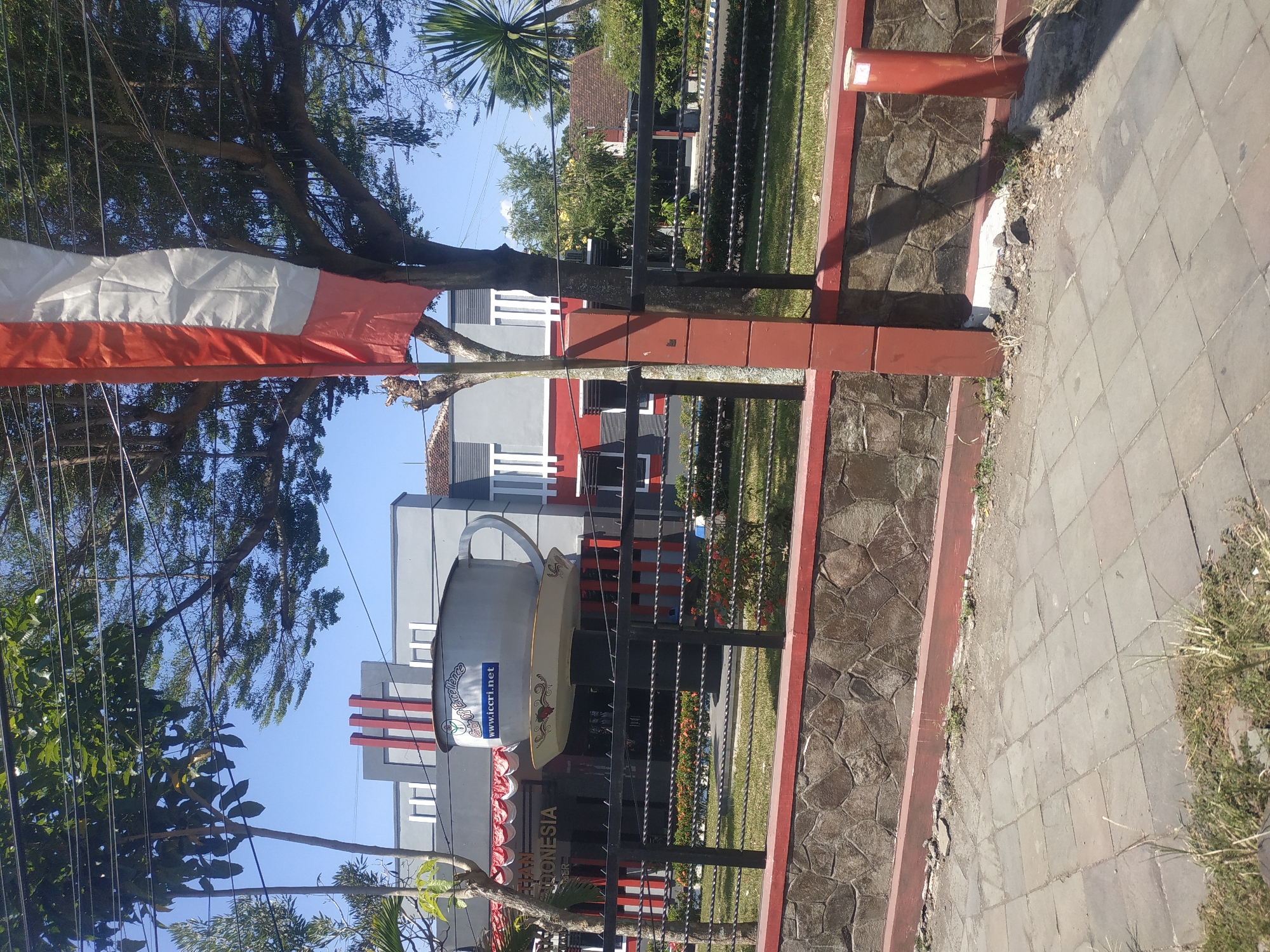 The image shows a large, decorative teacup sculpture perched on a pedestal in front of a building in Jember, Indonesia. The building features a modern architectural style with grey and red accents, and signage that reads "Pusat Unggulan Indonesia" and "www.ccri.net". A red and white Indonesian flag is visible to the right of the sculpture. The scene is set outdoors on a sunny day, with trees and greenery surrounding the area. A paved walkway is in the foreground, with a stone wall and fence separating the walkway from the building's grounds.