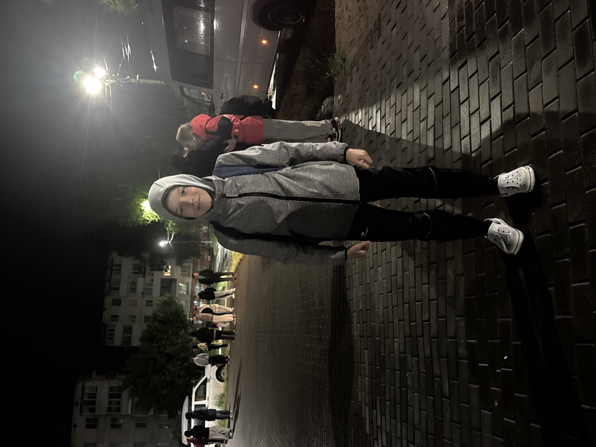 The image depicts an outdoor urban scene at night in the city of Chaikovsky, Russia. A young male, estimated to be a pre-teen, stands centrally in the foreground, facing the camera with a slight smile. He wears a grey zip-up hoodie with the hood drawn up, black athletic trousers with a white stripe at the knees, and white slip-on shoes. A blue backpack strap is visible over his right shoulder beneath his hoodie. His shadow is cast to his left.
In the mid-ground, a group of approximately ten individuals is gathered to the left, near two white vehicles parked partially on a grassy verge adjacent to a paved area. The dominant vehicle is a white passenger van, likely a Mercedes-Benz Sprinter, with the Cyrillic word "ЗАКАЗ" (ZAKAZ - 'hire' or 'order') visible on its side. Another white vehicle is partially obscured behind the group. The individuals are dressed in various casual attire, including jackets and hoodies; one person in a red jacket appears to be holding a small, light-colored dog. They are standing and conversing or waiting.
The background features two multi-story, light-colored apartment buildings typical of Soviet-era architecture, dimly lit. Trees are interspersed between the buildings and the mid-ground. The scene is illuminated by artificial light sources, prominently a bright streetlight with a visible glare positioned to the right of the main subject. The ground is paved with dark grey rectangular pavers, which exhibit wet, reflective patches, indicating recent precipitation. Areas of grass and dirt are visible alongside the paved surface where the vehicles are parked.