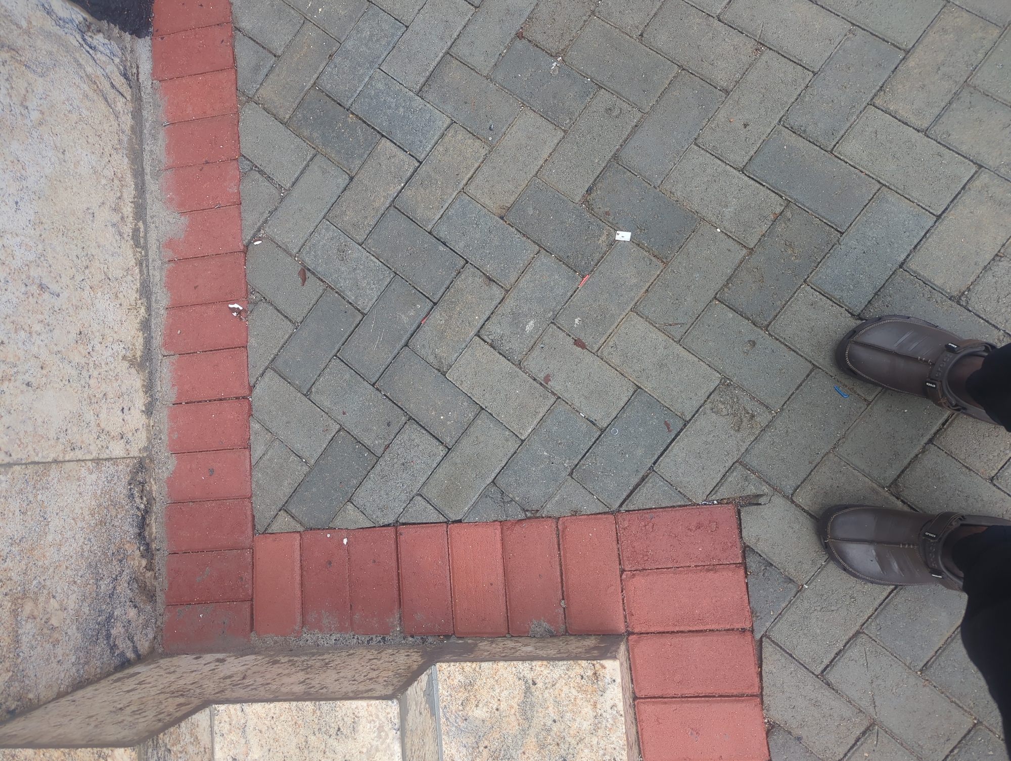 The image shows a close-up, overhead view of a paved walkway in Gwarinpa, Nigeria. The walkway is constructed with a border of red bricks laid horizontally along the edge. Inside the border, the majority of the walkway is paved with grey rectangular bricks arranged in a herringbone pattern. In the lower portion of the image, two feet are visible, wearing dark brown clogs. The feet are positioned on the grey brick paving. To the left of the red brick border, there are stone steps with a speckled texture. The scene appears to be an outdoor public space.