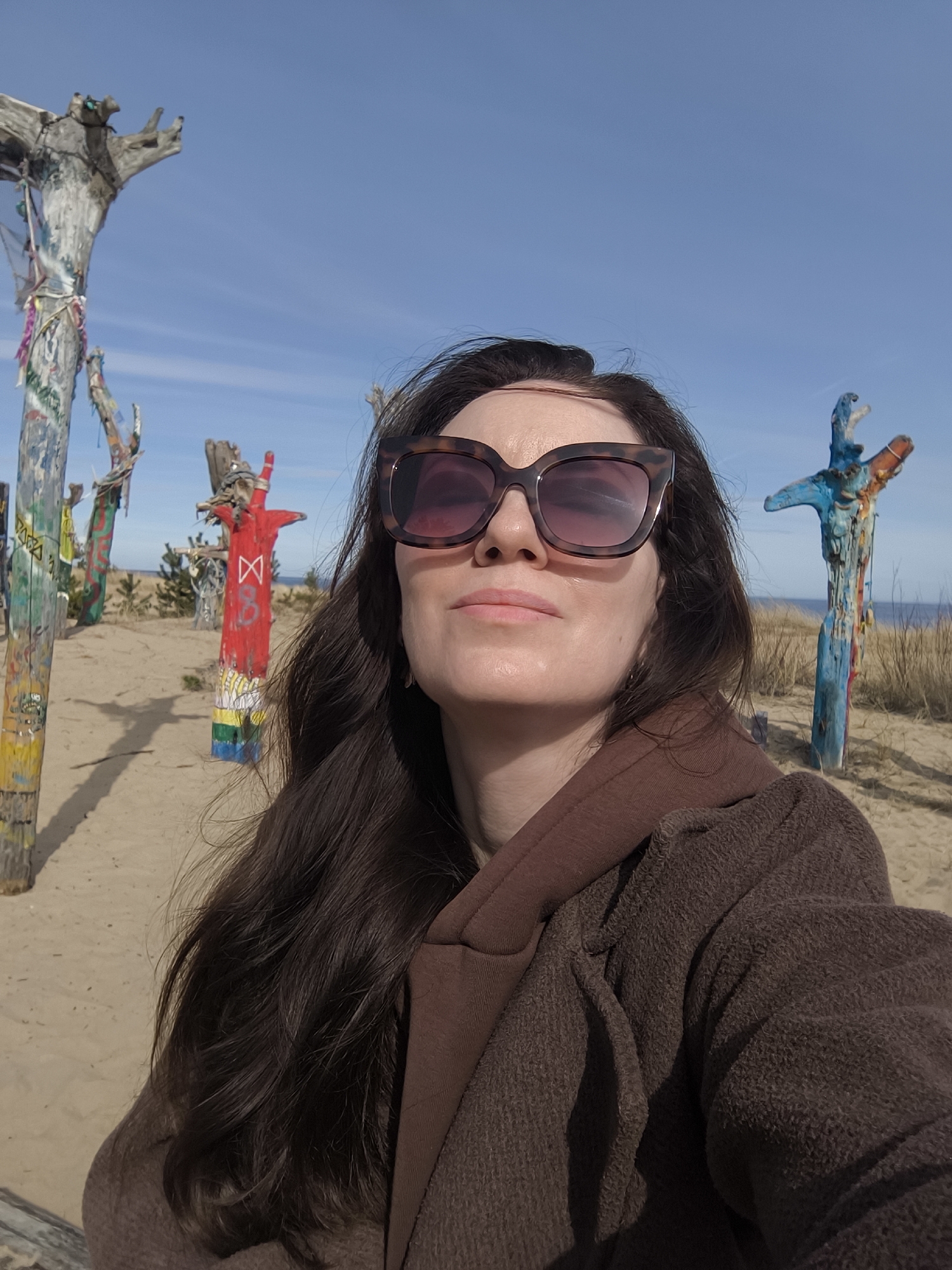 The image depicts a woman taking a selfie at the Gauja seaside nature reserve in Gauja, Latvia. The background consists of a sandy beach with multiple wooden totem-like structures of various colors and designs, and some vegetation. In the distance, there is a visible stretch of the sea under a blue sky with scattered clouds. The woman is wearing brown sunglasses and a brown layered jacket or coat. Her dark hair is long and flowing. The lighting suggests it is daytime.