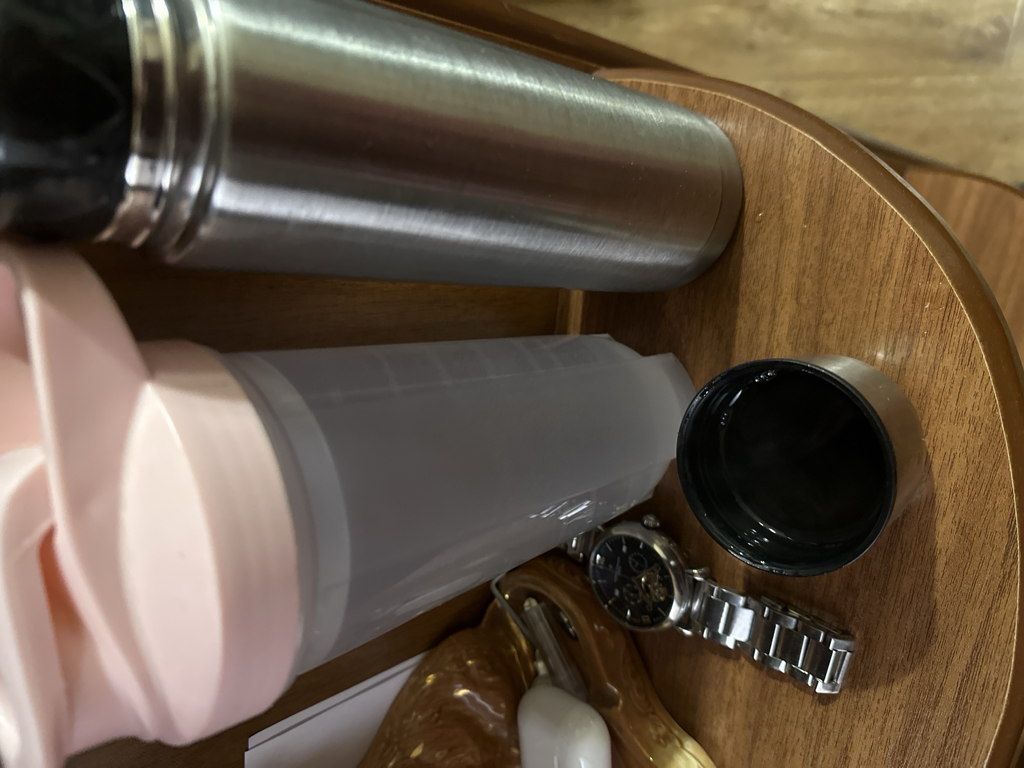 The image shows a close-up of several items on a wooden surface. There is a stainless steel water bottle laying horizontally across the top of the frame. Below it, there is a clear plastic shaker bottle with a pink lid. Next to the bottle is a black lid and a stainless steel watch. There is also a blurred figurine in the bottom of the image.