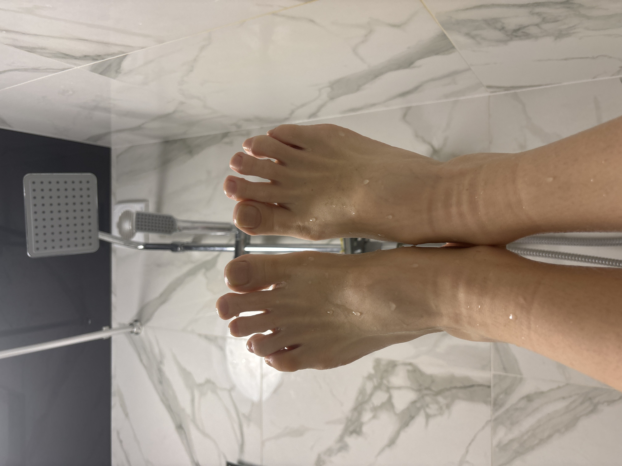 A pair of wet feet are positioned in a shower stall. The shower has a white and gray marble-patterned wall. A gray and chrome shower head and handle are present on the left side of the image. The scene is in an unidentified location; there is no forensic evidence to suggest the city of Sinyavino, Russia.