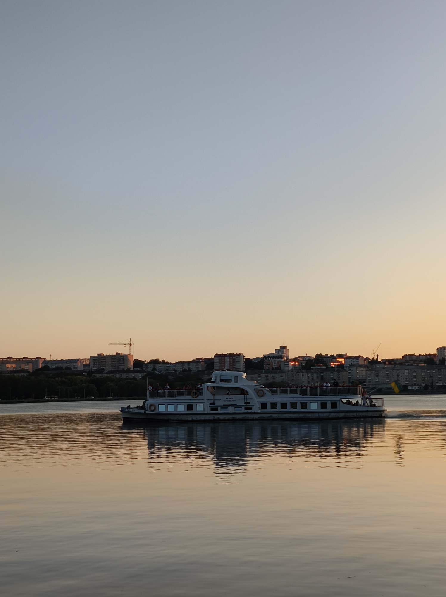 This is a full shot image capturing a sunset over the Ternopil Lake in Ternopil, Ukraine. The sky transitions from a pale blue at the top to a warm orange near the horizon. A passenger boat is depicted in the mid-ground, traversing the calm water. The boat, which appears to be a multi-deck vessel, has several passengers visible on its upper deck. Behind the boat, the Ternopil skyline is visible, characterized by a cluster of buildings and a prominent construction crane. The water reflects the colors of the sky and the boat, creating a serene and atmospheric scene. The water is calm with gentle ripples.