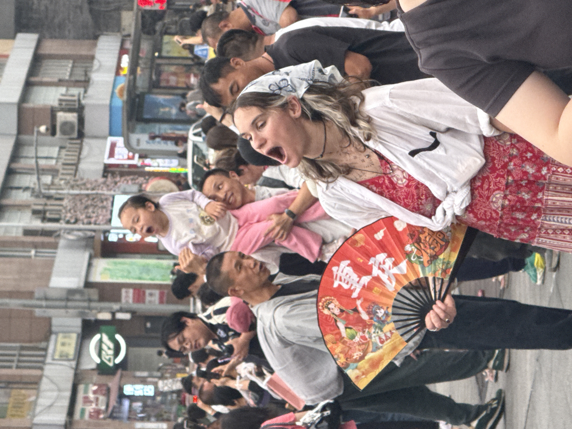 The image shows a crowd of people, possibly tourists, in an urban setting in Yuzhong District, China. The group is tightly packed. People are wearing casual clothing. A woman in the foreground is shouting with her mouth open, wearing a floral dress and a white shirt. A man in front of her is holding a fan with traditional Chinese illustrations. The background shows buildings with signage, advertisements, and air conditioning units, suggesting a commercial area.