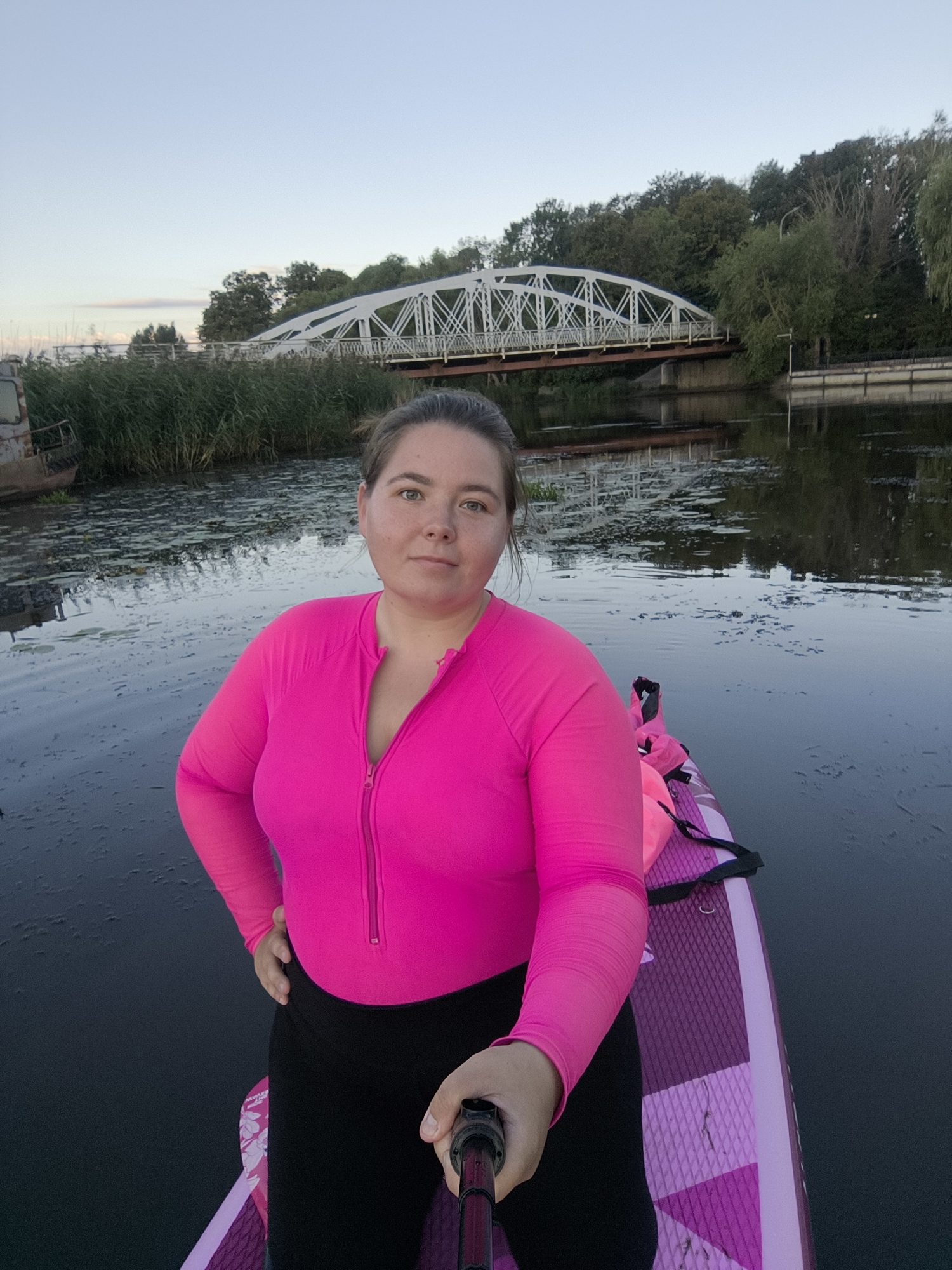 In Ushakovo, Russia, a woman is pictured on a stand-up paddleboard on a body of water. She is wearing a bright pink long-sleeved rash guard and black leggings. Her hair is pulled back. The paddleboard is purple and pink. In the background, a white truss bridge spans the water. Reeds line the shore, and trees are visible beyond the bridge. The sky is clear with a hint of sunset colors.