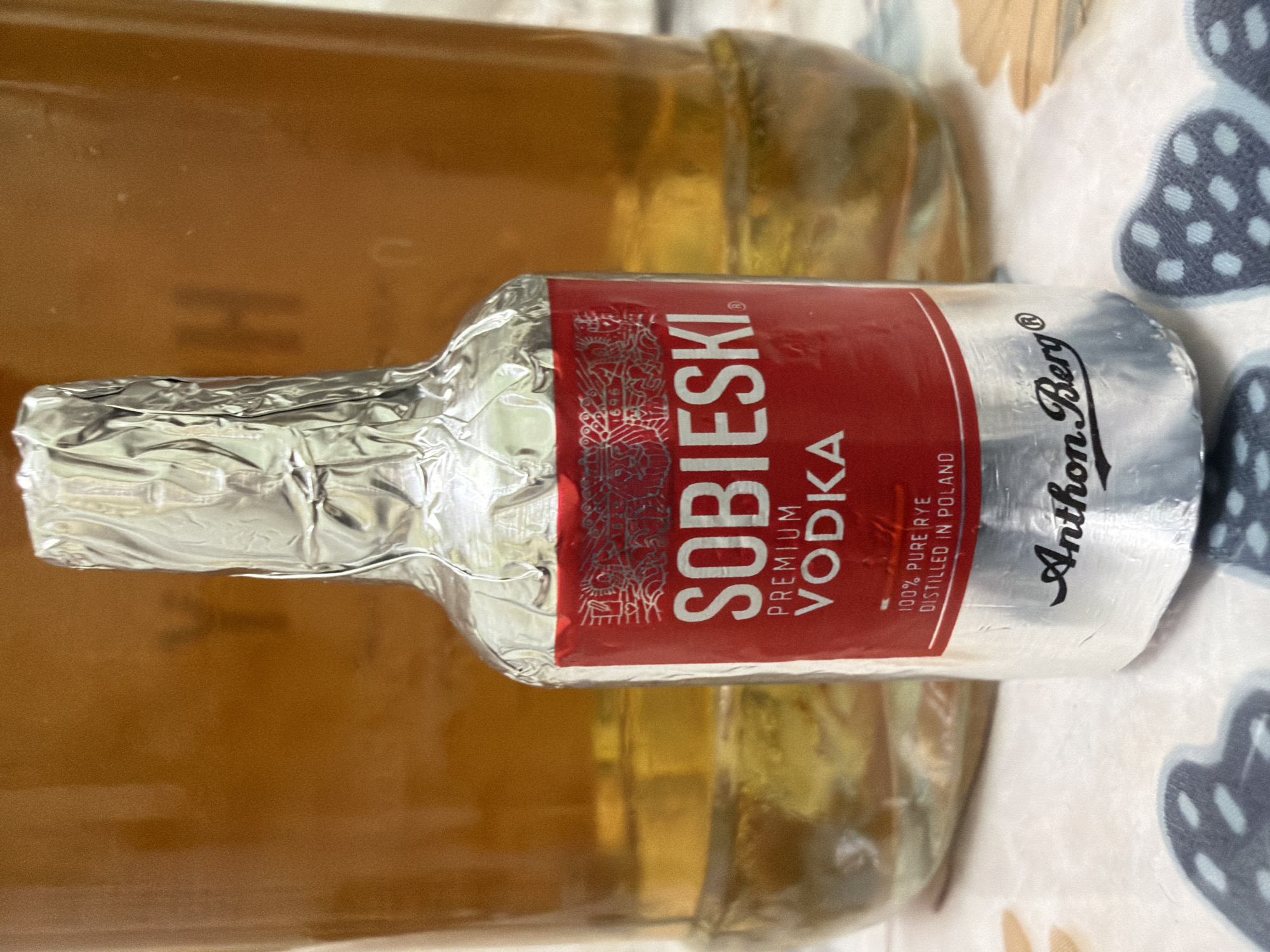 An Anthon Berg chocolate liqueur bottle, wrapped in silver foil, stands in front of a larger bottle containing a light brown liquid. The Anthon Berg bottle has a red label that reads "SOBIESKI PREMIUM VODKA" and "100% PURE RYE DISTILLED IN POLAND". The larger bottle, in the background, is a glass bottle with a golden-amber liquid. The scene is set on a table covered with a patterned tablecloth. The lighting is ambient. No people or actions are depicted. The image is a close-up shot of the bottles. There is no information available to determine the specific location in Kyiv, Ukraine.