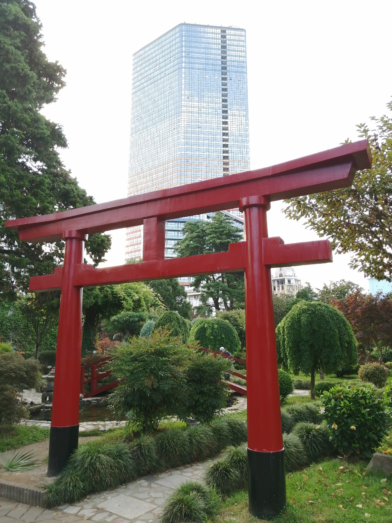 The image depicts a red torii gate in the foreground, a prominent Japanese architectural feature. Behind the gate, a meticulously maintained Japanese garden is visible, complete with lush greenery, a small pond, and a red wooden bridge. In the background, a modern, glass-covered skyscraper pierces the sky, suggesting an urban setting. This juxtaposition of traditional Japanese elements with contemporary architecture strongly indicates the location is the Batumi Japanese Garden in Batumi, Georgia. A person in a white hard hat is seen on the red bridge, likely a visitor or worker. The scene is characterized by a mix of natural and man-made structures, creating a unique visual contrast.