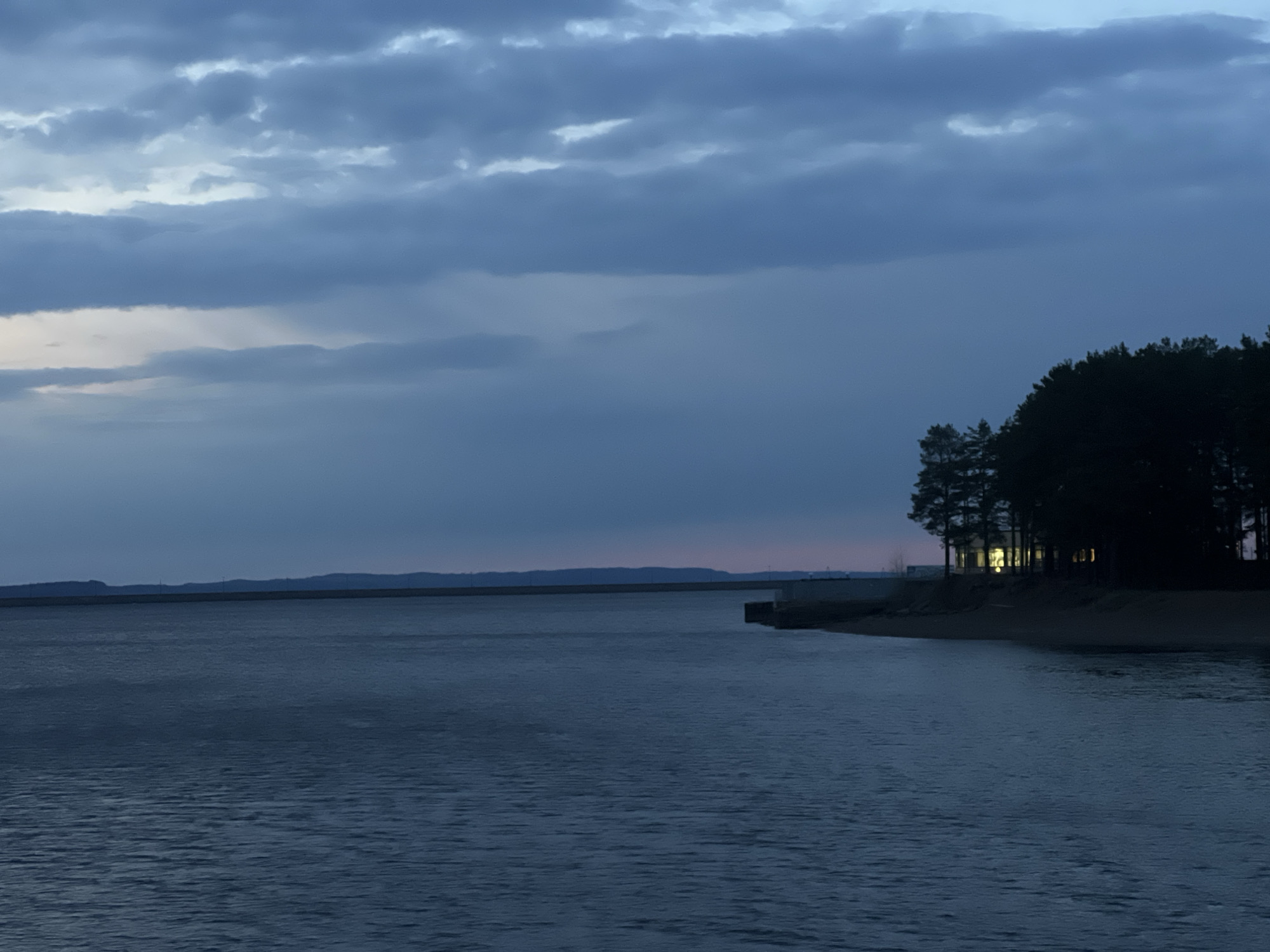 A wide shot captures a tranquil twilight scene over a large body of water, likely a reservoir, in Chaikovsky, Russia. The sky is largely covered by heavy, dark blue-gray clouds, with lighter patches showing through, hinting at the last vestiges of daylight or the first light of dawn.
The expansive water body occupies the majority of the lower frame, its surface reflecting the subdued light with gentle ripples. A low, distant horizon line, possibly a dam wall or landmass, stretches across the water in the background. On the right, a dense line of tall, dark coniferous trees forms a prominent silhouette against the dim sky. Nestled within these trees, a building or structure is visible, with multiple windows casting a warm, bright yellow light, providing a stark contrast to the cool, somber tones of the surrounding landscape. A small concrete structure, resembling a pier or part of a shoreline fortification, extends a short distance into the water from the tree-lined bank.
The time of day is clearly dusk or dawn, characterized by the low light conditions and the artificial illumination from the building. The weather appears overcast. No people or visible text are present in the image. The scene evokes a sense of quiet serenity.