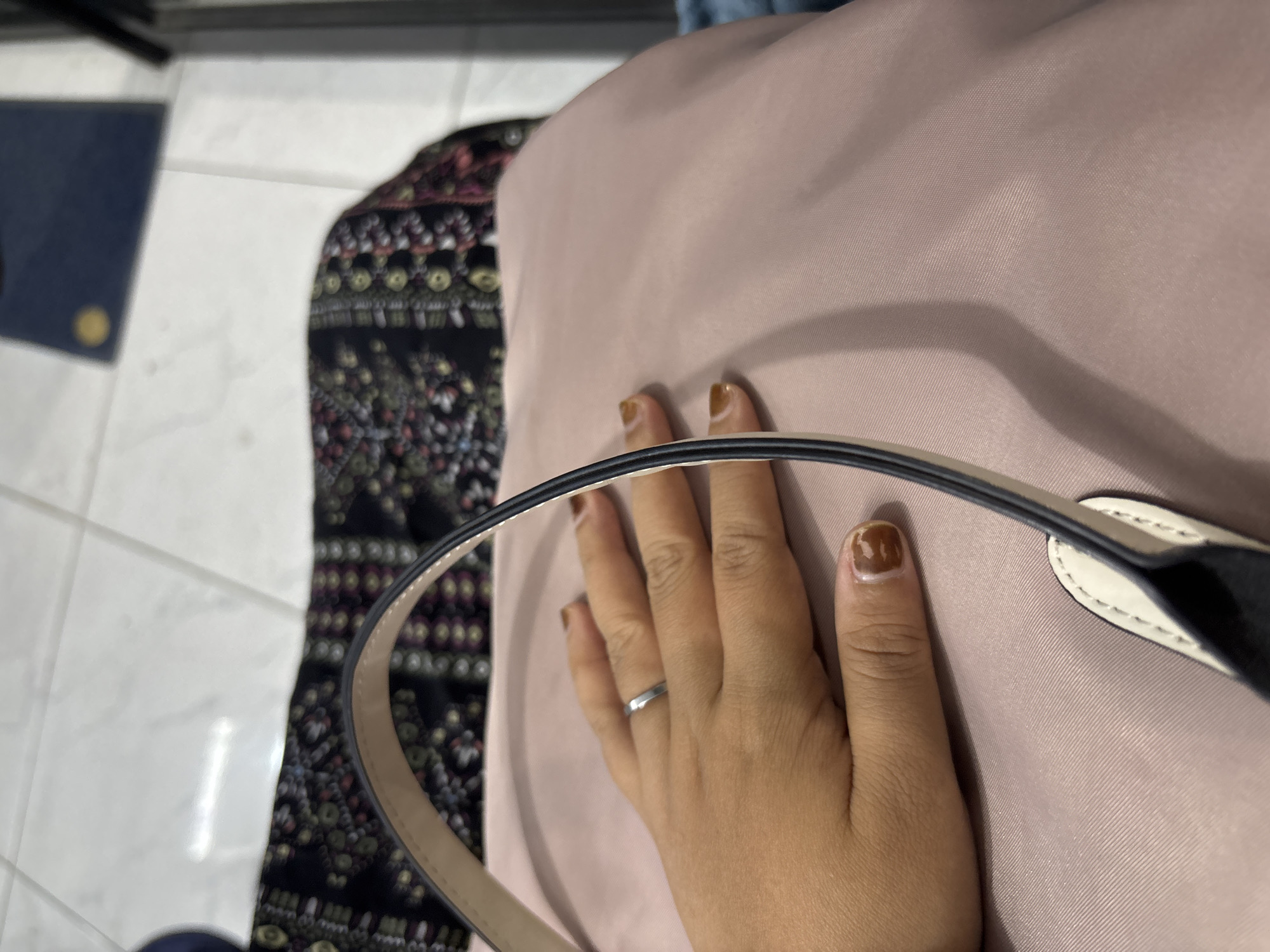 A close-up, top-down view shows a person's hand resting on a pink fabric surface, likely a bag. The hand has light brown nail polish, with some chips visible on the thumb. A thin, silver ring is worn on the ring finger. Behind the hand and fabric is a dark fabric with a colorful, intricate pattern. The background consists of light-colored floor tiles. No specific location within Wanajaya, Indonesia is discernible from the image.