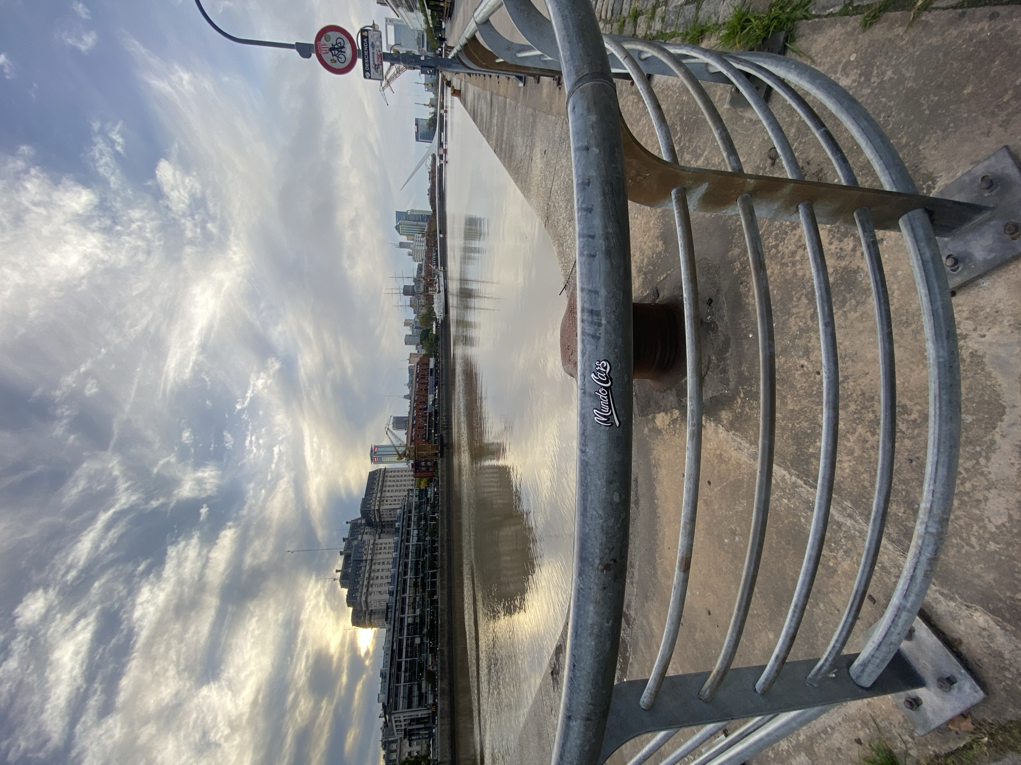 This image, likely taken in Buenos Aires, Argentina, shows a waterfront scene along the Río de la Plata. The foreground features a metal railing with a sticker that reads "Mundo Cars." Beyond the railing is a concrete walkway and then water. The water reflects the city skyline, complete with buildings, cranes, and the sunset. The sky is partially cloudy, with the sun creating a bright glow on the horizon. A traffic sign indicating bicycle restrictions is visible.
