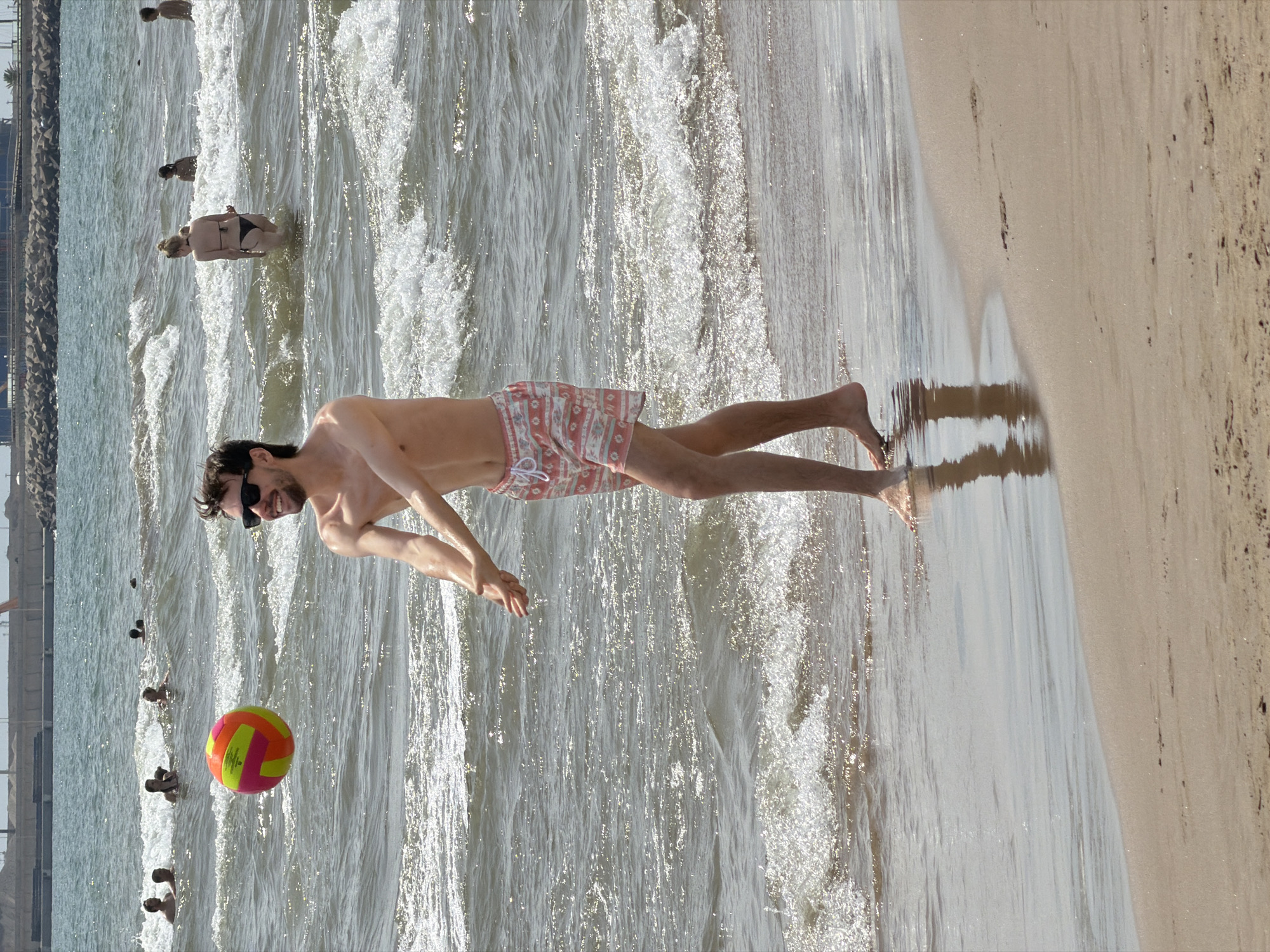 The image shows a group of people at a beach on a sunny day. In the foreground, a man is lying on his back in shallow water, wearing patterned swim shorts. He has sunglasses on and is smiling. A colorful beach ball is floating near him. In the background, other people are visible in the water, some swimming or wading. The scene appears to be a recreational setting at the beach, with the ocean and sand clearly depicted. The lighting suggests it is daytime, and the bright sun creates sparkling reflections on the water. The overall atmosphere seems relaxed and enjoyable, with people engaging in typical beach activities. There is no visible text in the image.