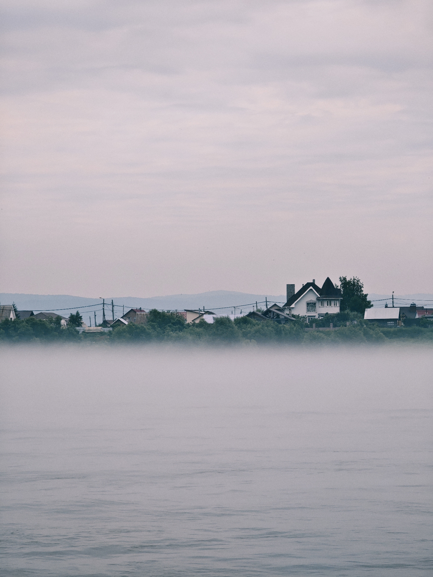 The image depicts a foggy morning scene on the Yenisei River in Zheleznogorsk, Russia. In the foreground, the calm river surface is visible, with subtle ripples indicating a gentle current. A thick layer of fog hovers low over the water, obscuring the lower portion of the distant riverbank. Beyond the fog, a cluster of houses and trees forms a village silhouette against a hazy sky. The architecture of the houses appears to be typical of a rural Russian settlement. Power lines and poles are also visible, suggesting the presence of infrastructure. The sky is overcast with soft, diffused light, contributing to the muted and serene atmosphere of the scene.