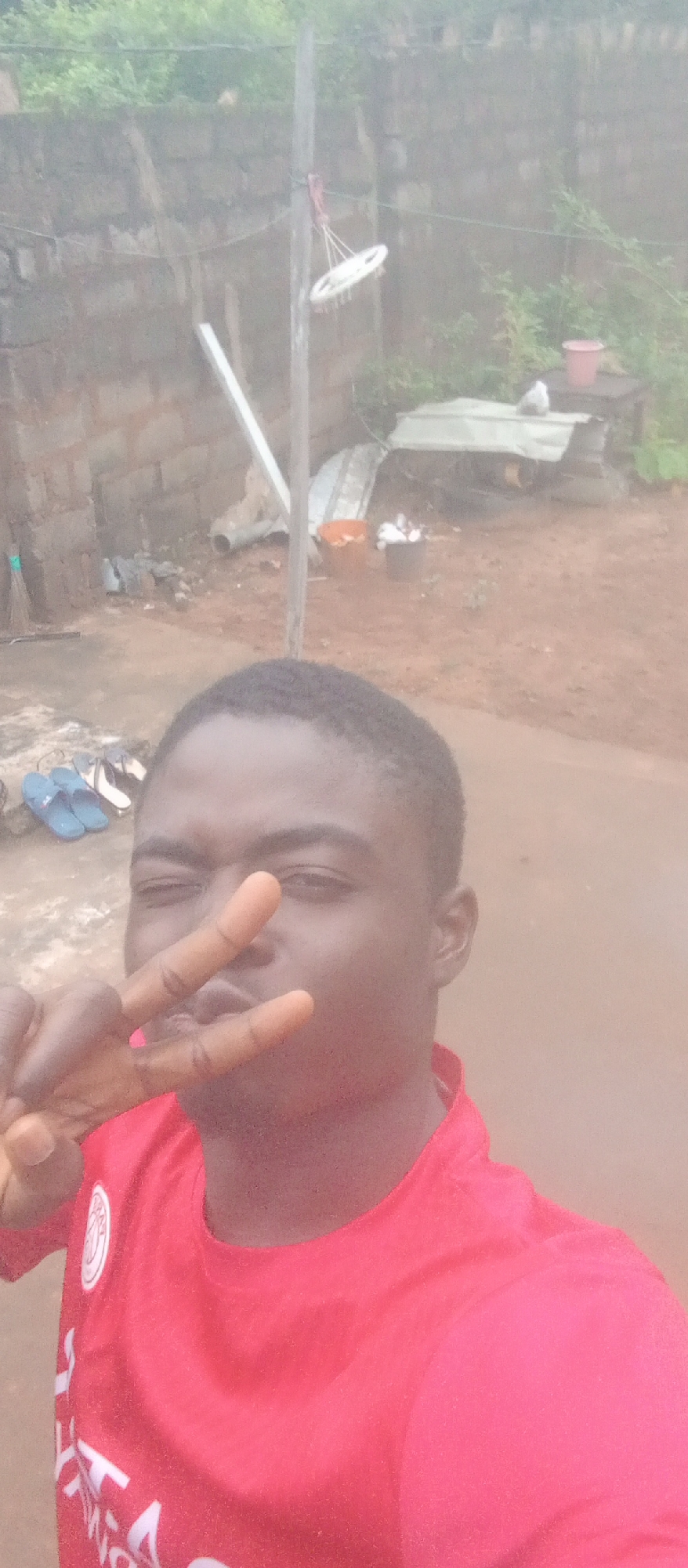 The image shows a young Black male in the foreground, taking a selfie. He is wearing a red t-shirt with a white emblem on the left chest. He is making a peace sign with his right hand, with his index and middle fingers extended towards the camera. His left hand is partially visible. He is looking at the camera with his eyes partially closed.
In the background, there is a brick wall, a dirt ground, and some greenery. To the left of the male, there are a pair of blue flip-flops and a pair of white sandals on the ground. In the middle ground, there is a wooden post with a cleaning brush hanging from it by strings. There are also some buckets and other miscellaneous items in the background, including what appears to be a corrugated metal roof. The overall environment appears to be an outdoor space, possibly a yard or alleyway, with a somewhat cluttered and unkempt appearance. The lighting suggests it may be daytime, but the sky is overcast, giving the image a somewhat muted and hazy quality. The setting is consistent with a residential area in Ogidi, Nigeria.
