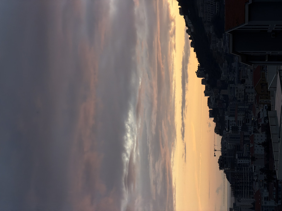 A sideways view showcases an urban landscape against a sunset sky. Buildings stretch along the horizon, their details obscured by shadows. The sky transitions from a dark, cloudy gray to a vibrant golden hue near the horizon, where the sun's light casts a warm glow. A construction crane is visible amidst the buildings, adding to the urban setting.