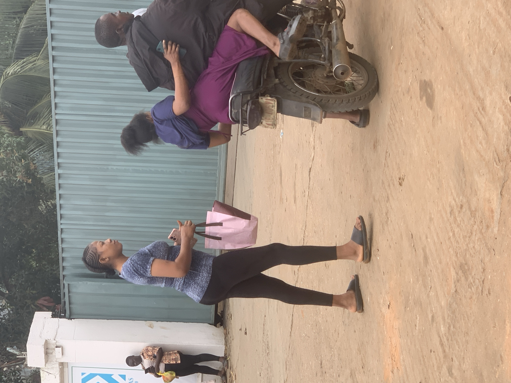 In the city of Shagamu, Nigeria, a street scene unfolds with several individuals and a motorcycle. A woman, wearing a blue patterned t-shirt and black leggings, stands on a dirt path, holding a pink and white shopping bag and a mobile phone. Her hair is styled in braids. To her right, a woman in a purple dress and dark blue wrap is seated on the back of a motorcycle, holding onto the driver. The driver, wearing a black jacket, is partially visible. Another person, dressed in a plaid shirt and black pants, stands near a white pillar and a gate, holding a mobile phone and a yellow bag. The background features a light blue corrugated metal fence and green foliage, suggesting an outdoor urban environment.